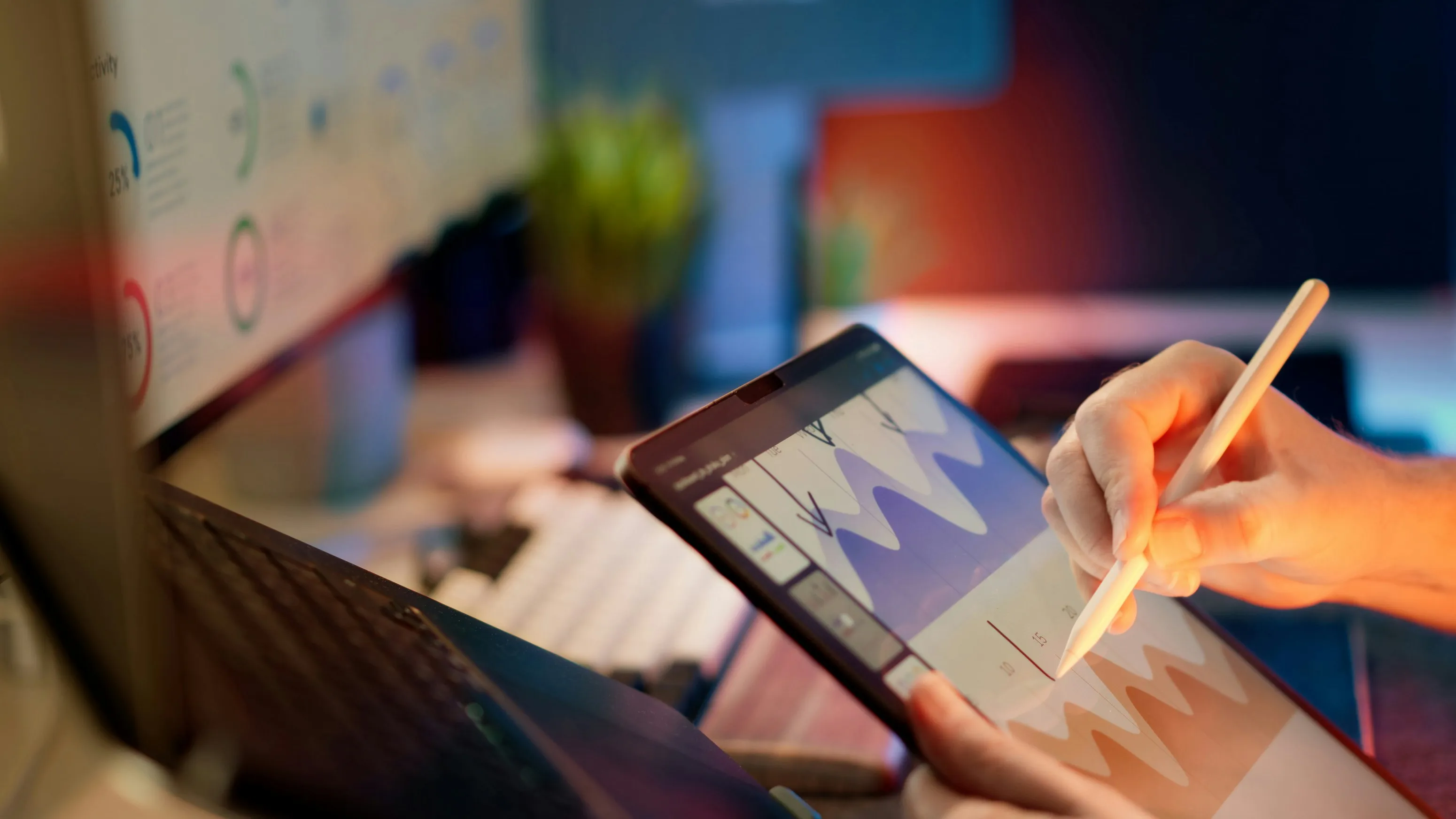 Person using a stylus on a tablet displaying colorful graph charts with a laptop and blurred monitors in the background.