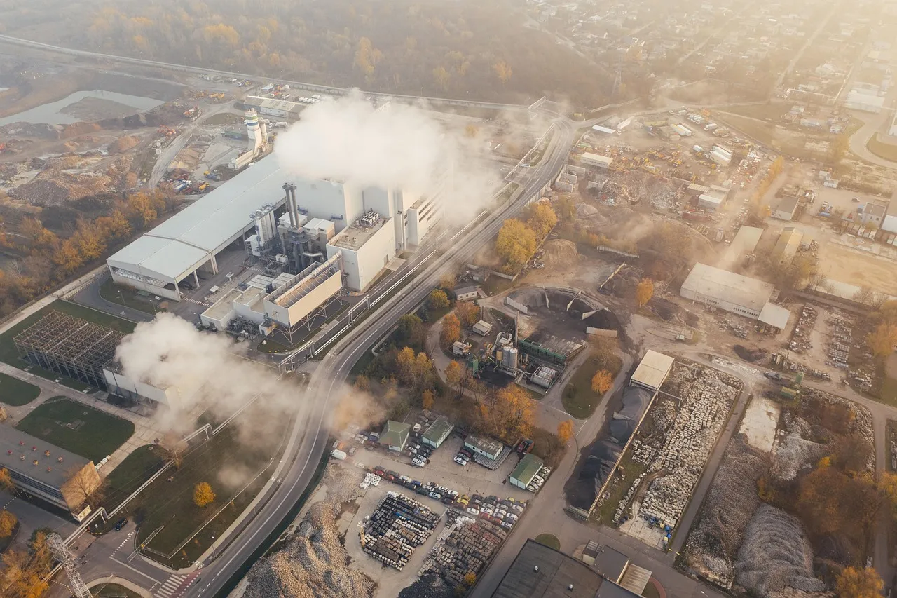 Aerial view of an industrial plant emitting smoke with surrounding roads, buildings, and storage areas under soft sunlight.