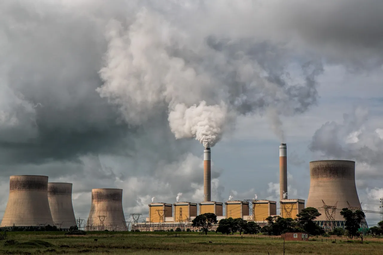 Industrial power plant with large cooling towers and smokestacks emitting thick white and gray smoke under a cloudy sky.