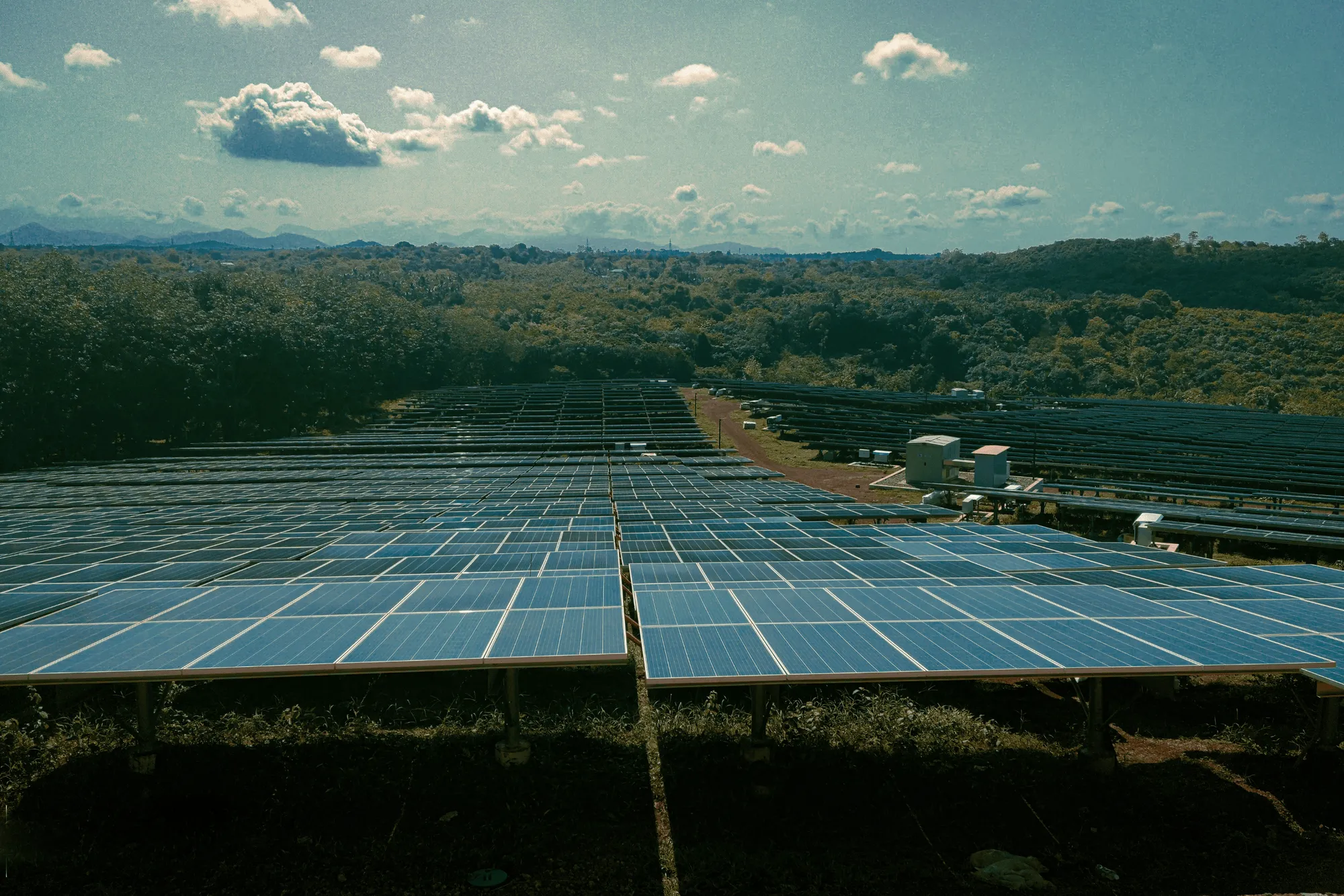 Large solar panel farm with rows of panels under a partly cloudy sky, surrounded by green hills and trees.