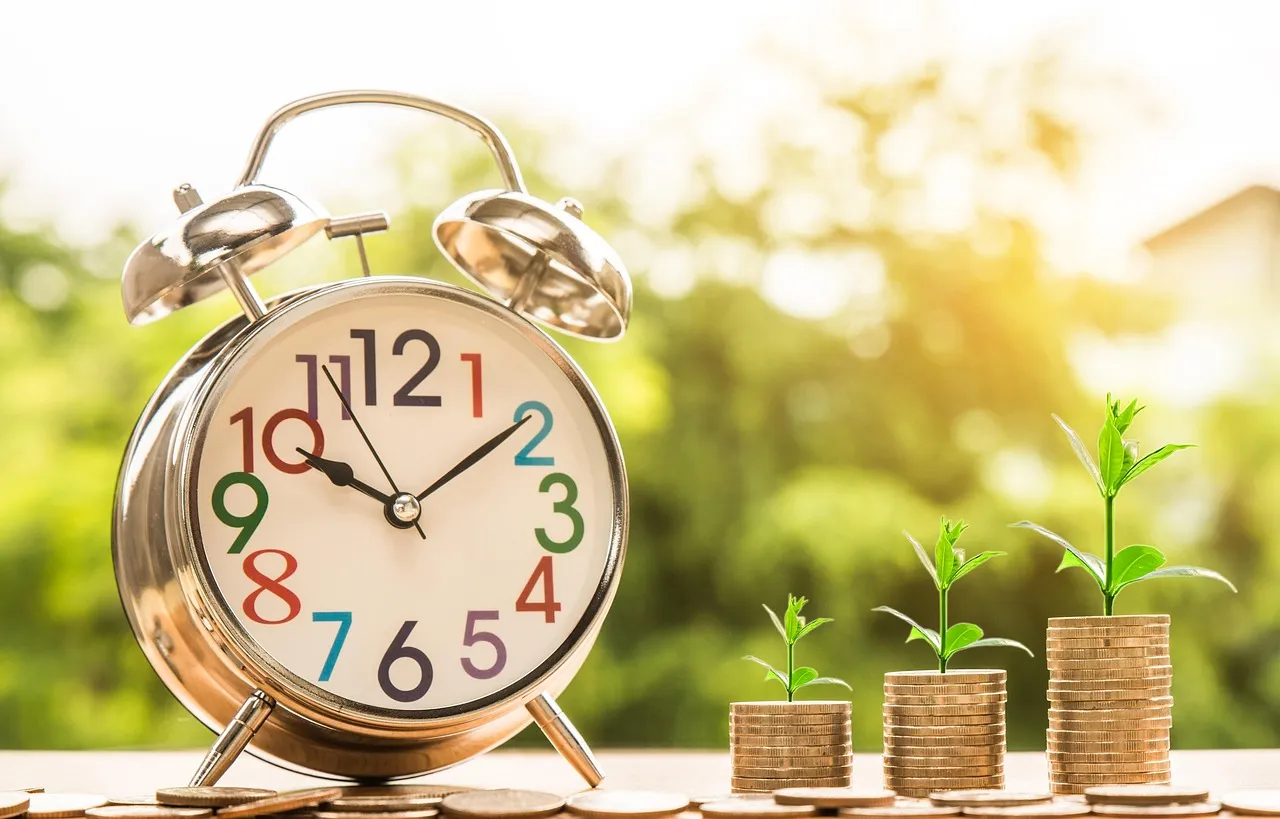 Metallic alarm clock with colorful numbers beside stacks of coins with growing green plants, symbolizing growth over time.