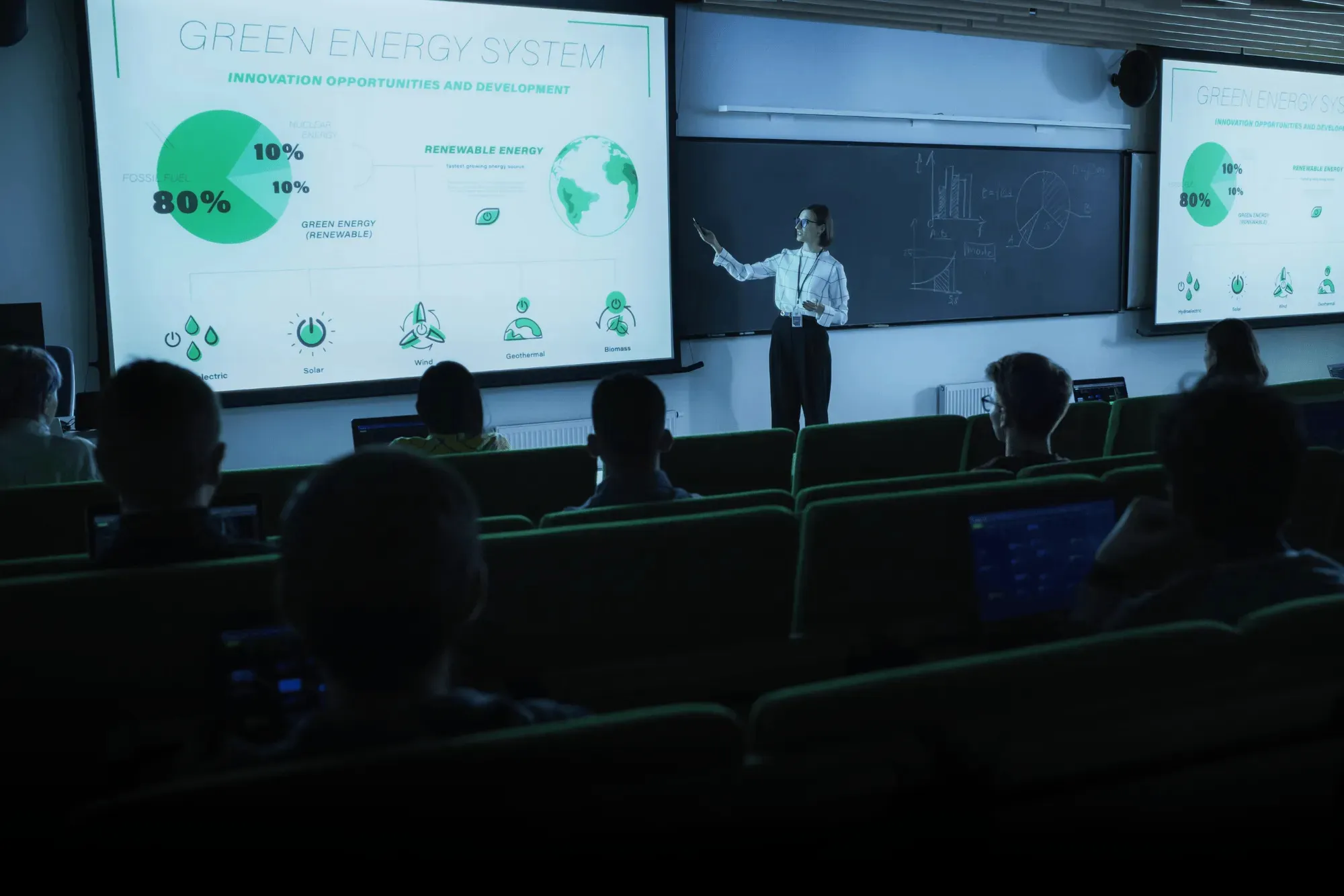 A woman giving a presentation on green energy systems to an audience seated in a dark classroom.