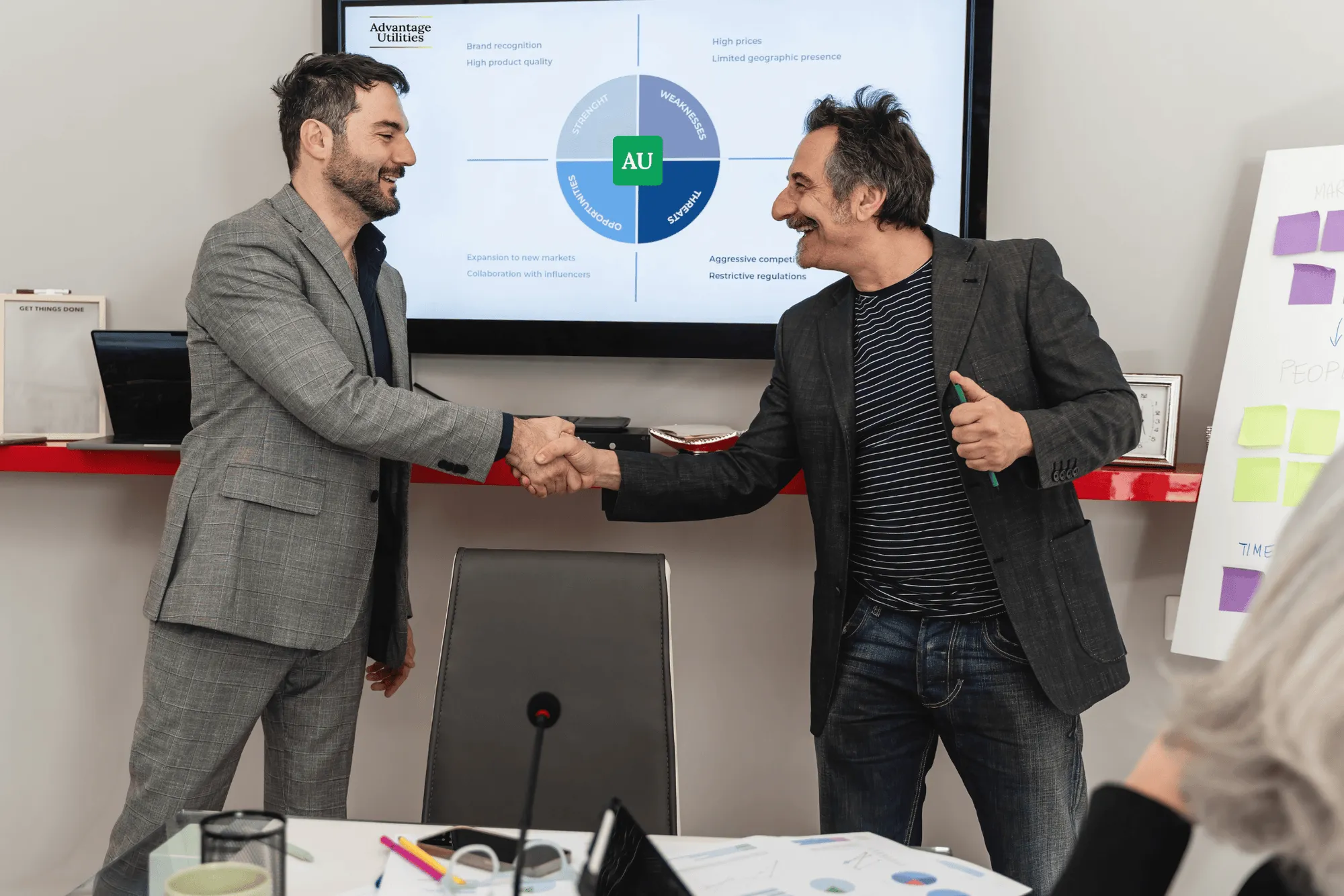 Two businessmen shaking hands in a meeting room with a presentation screen and a whiteboard with sticky notes in the background.