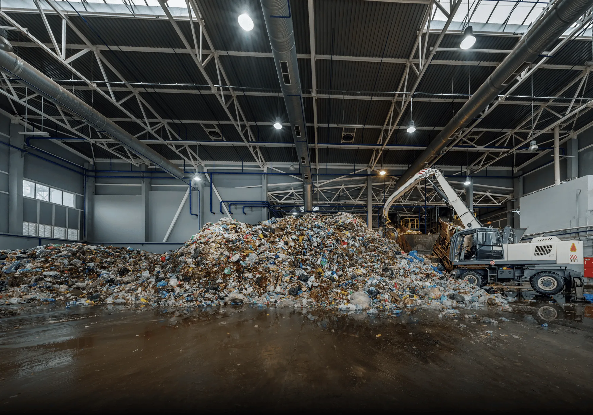 Large indoor waste processing facility with a tall pile of mixed garbage and an excavator sorting the trash.