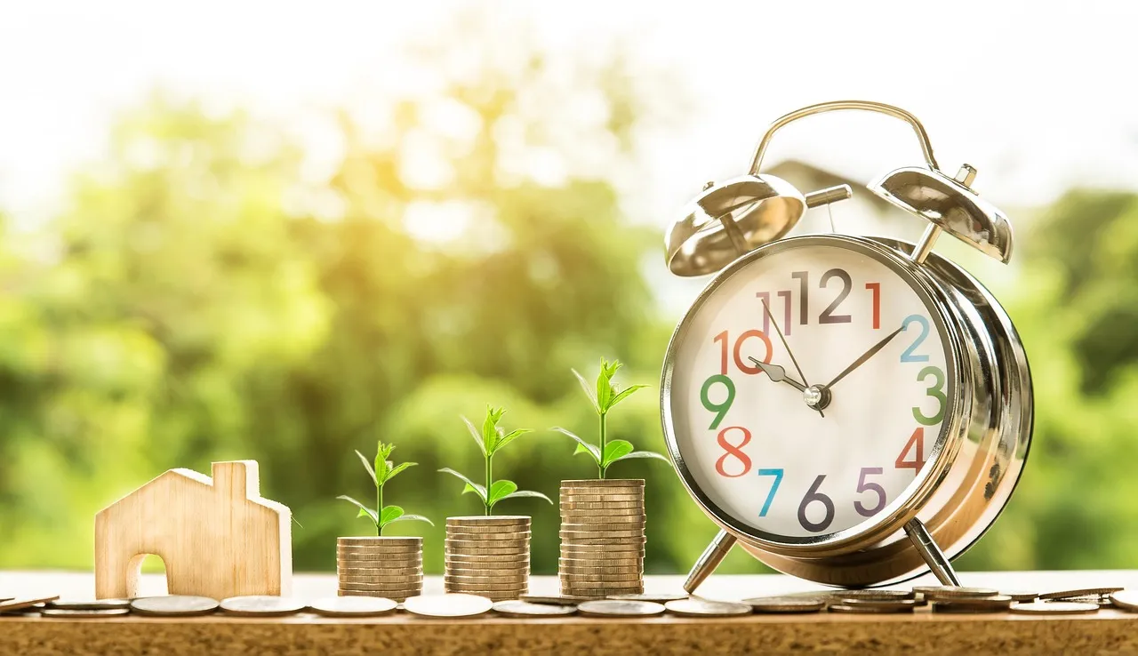 Wooden house model with stacks of coins growing in height topped with small green plants, next to a colorful alarm clock.