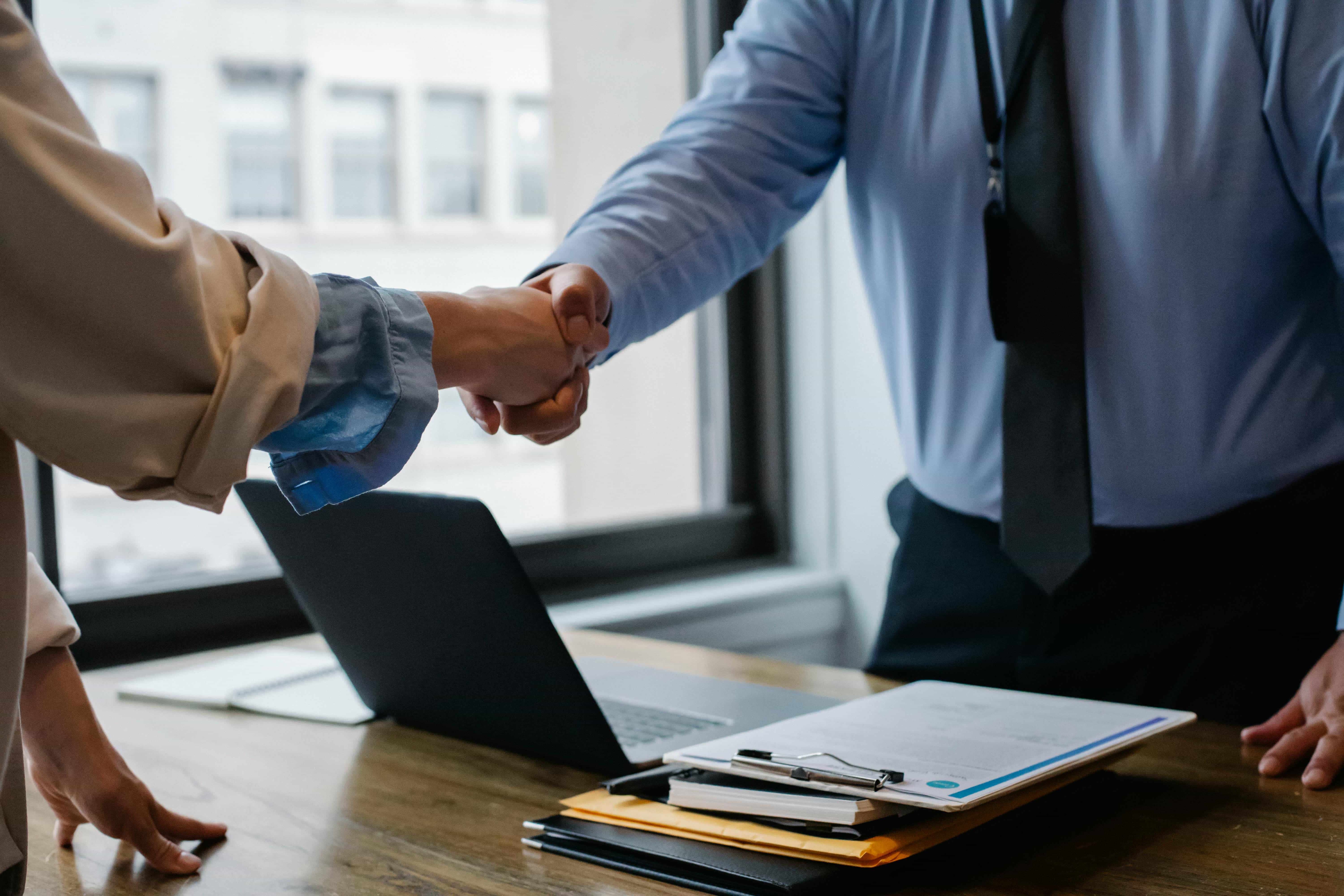 Two people shaking hands over a desk with a laptop and documents, symbolizing a business agreement.