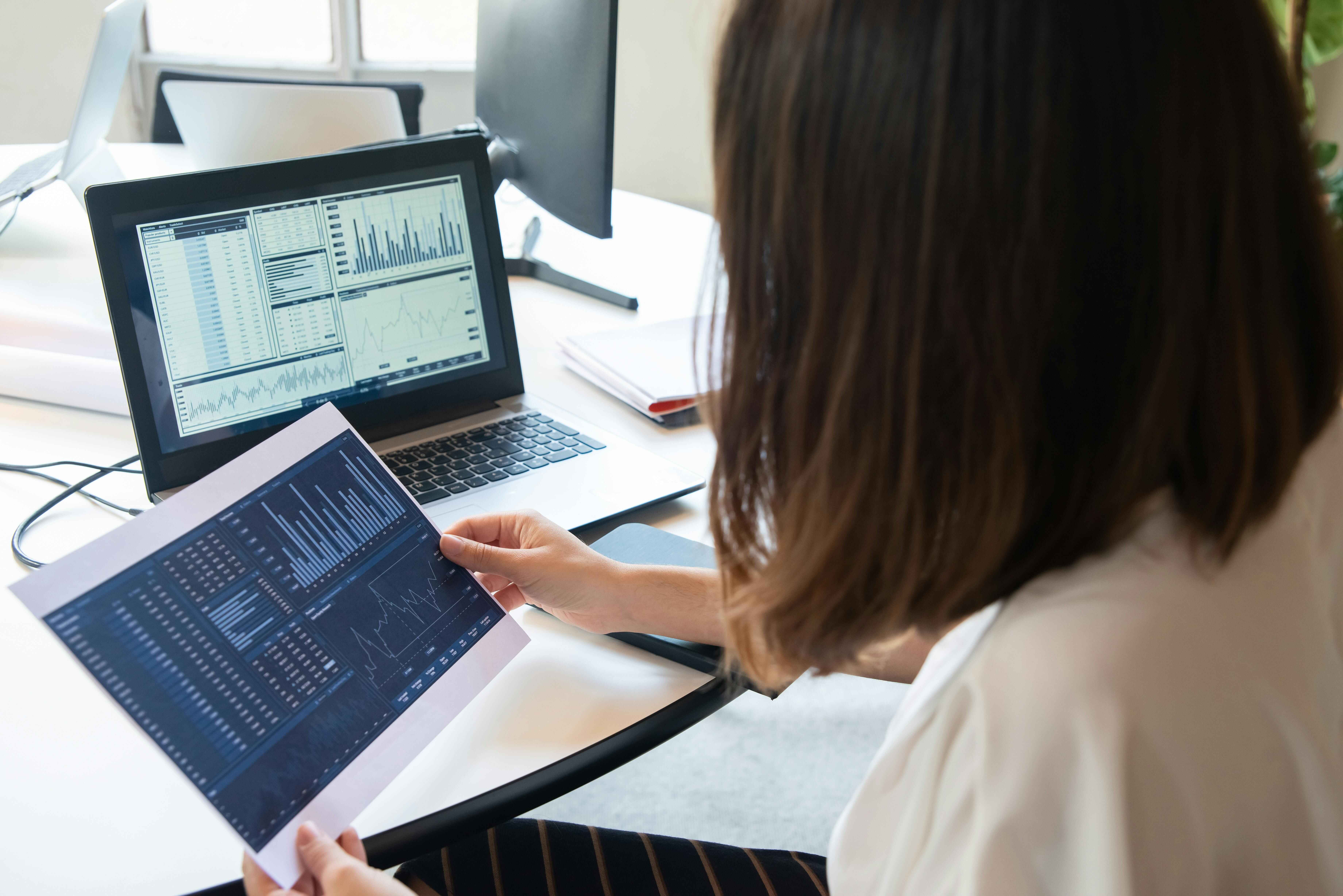 Person reviewing printed data charts and analytics graphs with similar information displayed on a laptop screen on a desk.