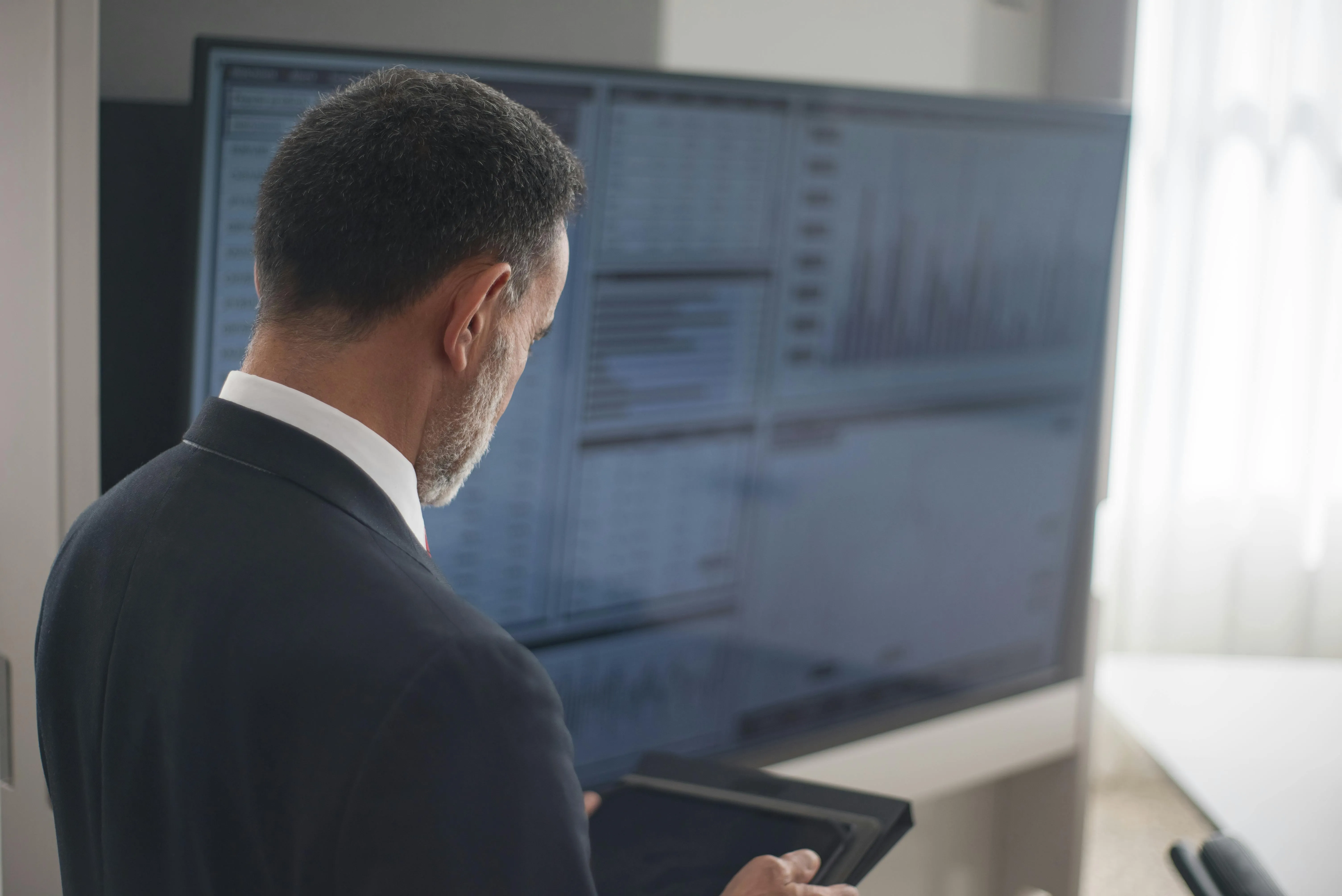 Man in a suit looking at a tablet with a large screen displaying charts and data in the background.