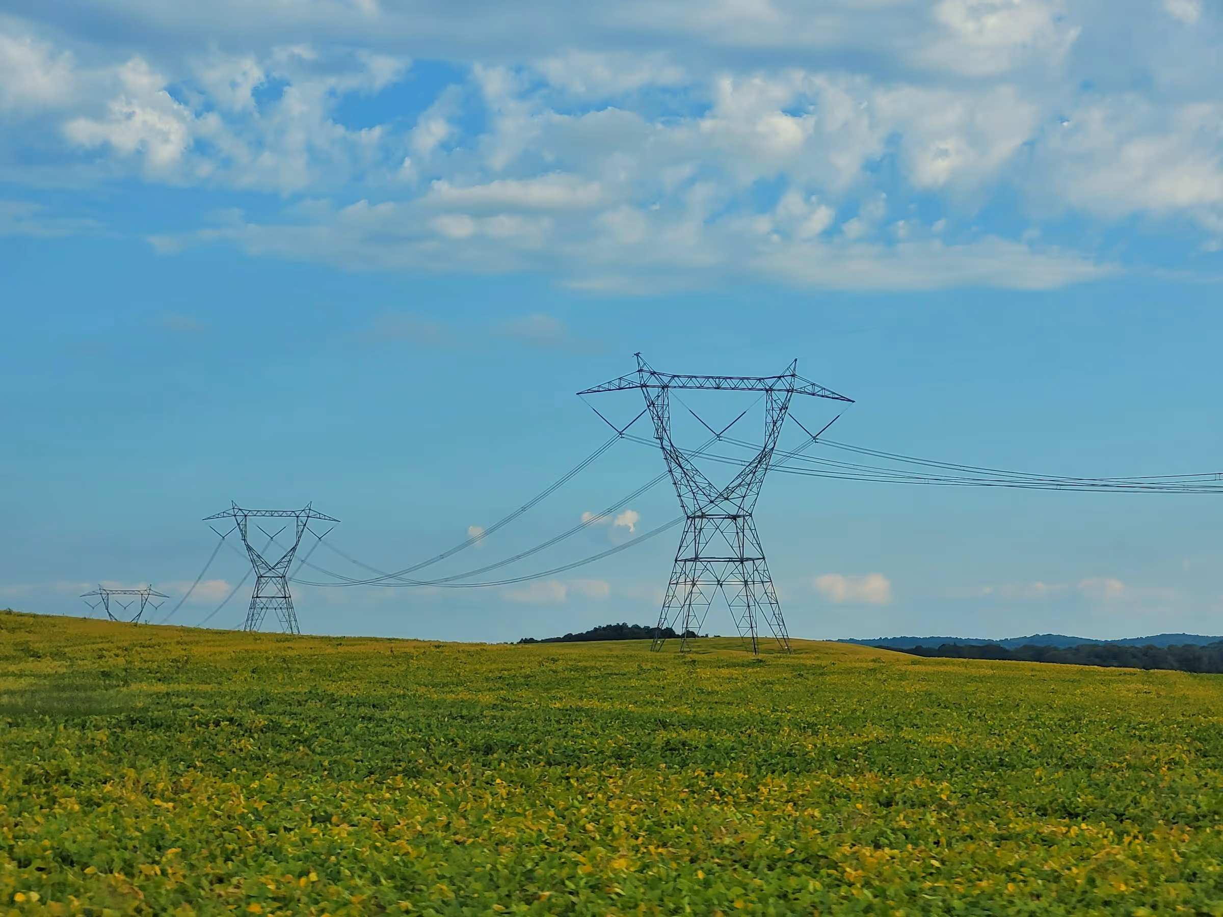 High-voltage power lines supported by steel lattice towers over a green field under a partly cloudy sky.