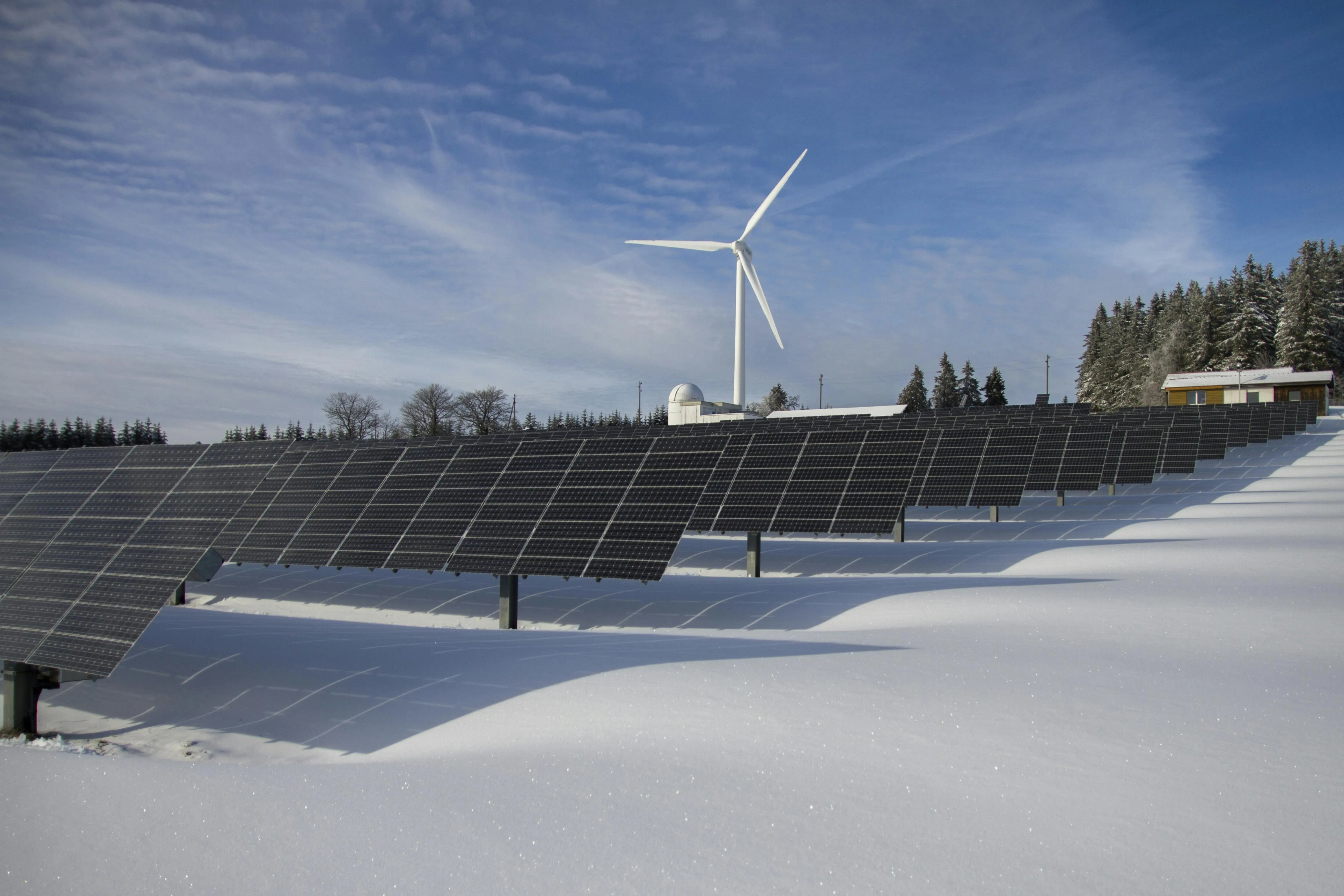 Rows of solar panels on snow-covered ground with a wind turbine and trees in the background under a partly cloudy sky.