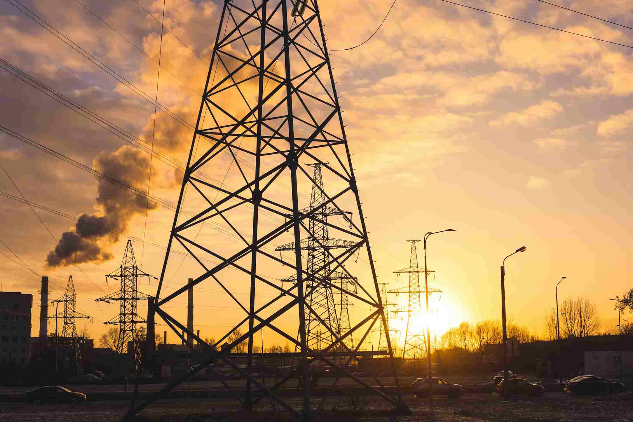 Silhouettes of electrical transmission towers against a sunset sky with smoke rising from an industrial chimney.