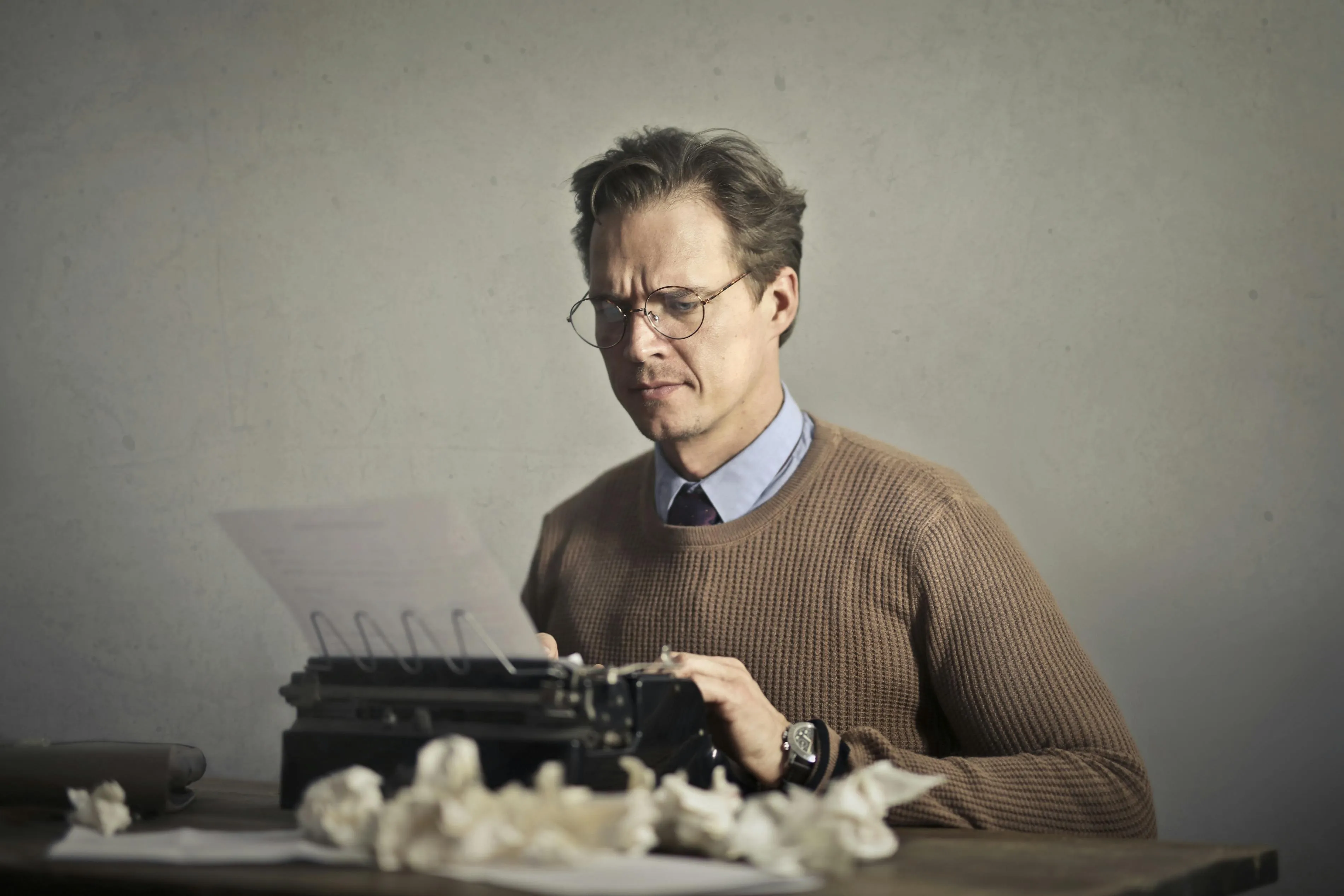 Man in glasses and brown sweater typing on a typewriter with crumpled papers on the table.