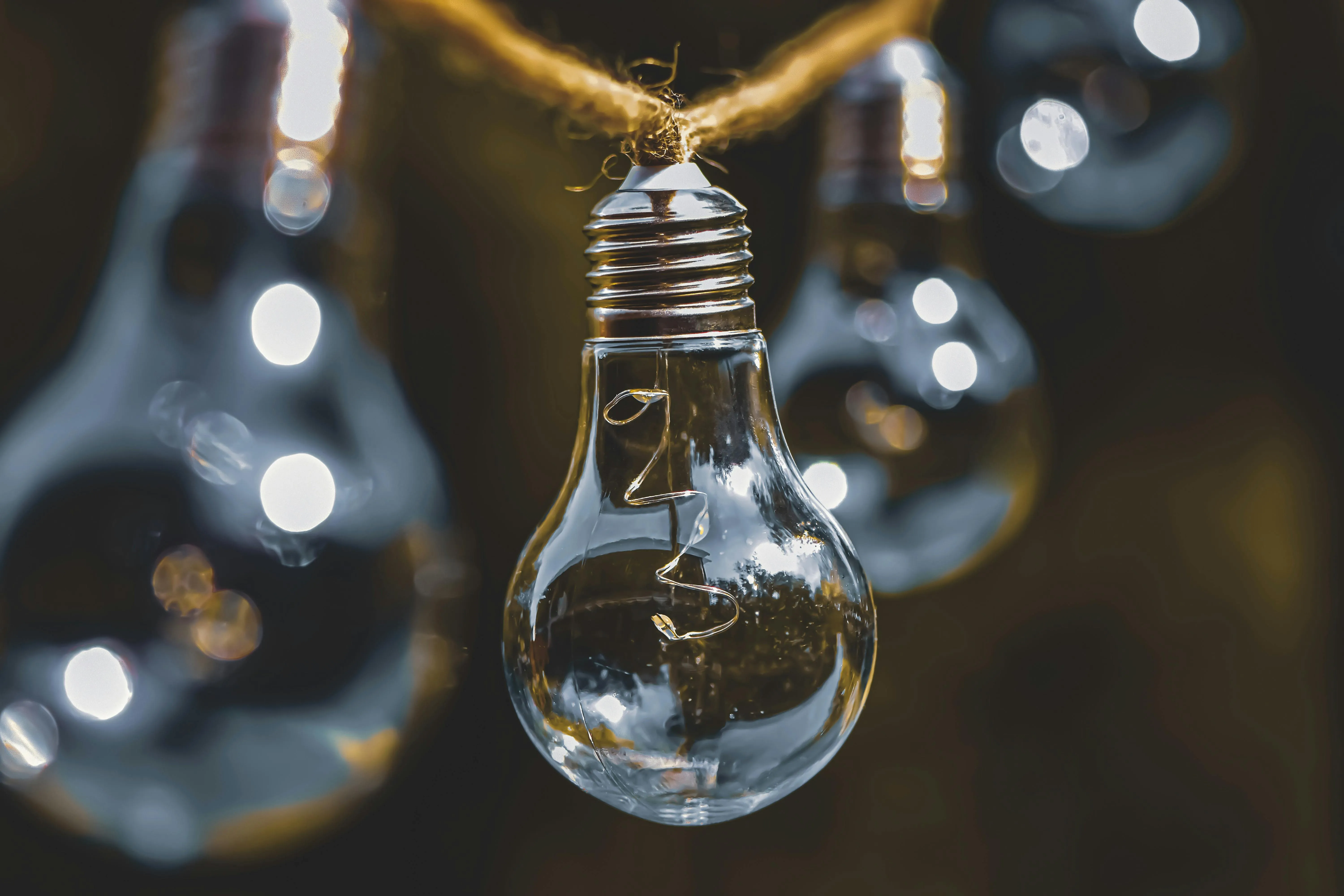 Close-up of a clear incandescent light bulb hanging from a brown rope with blurred light bulbs in the background.