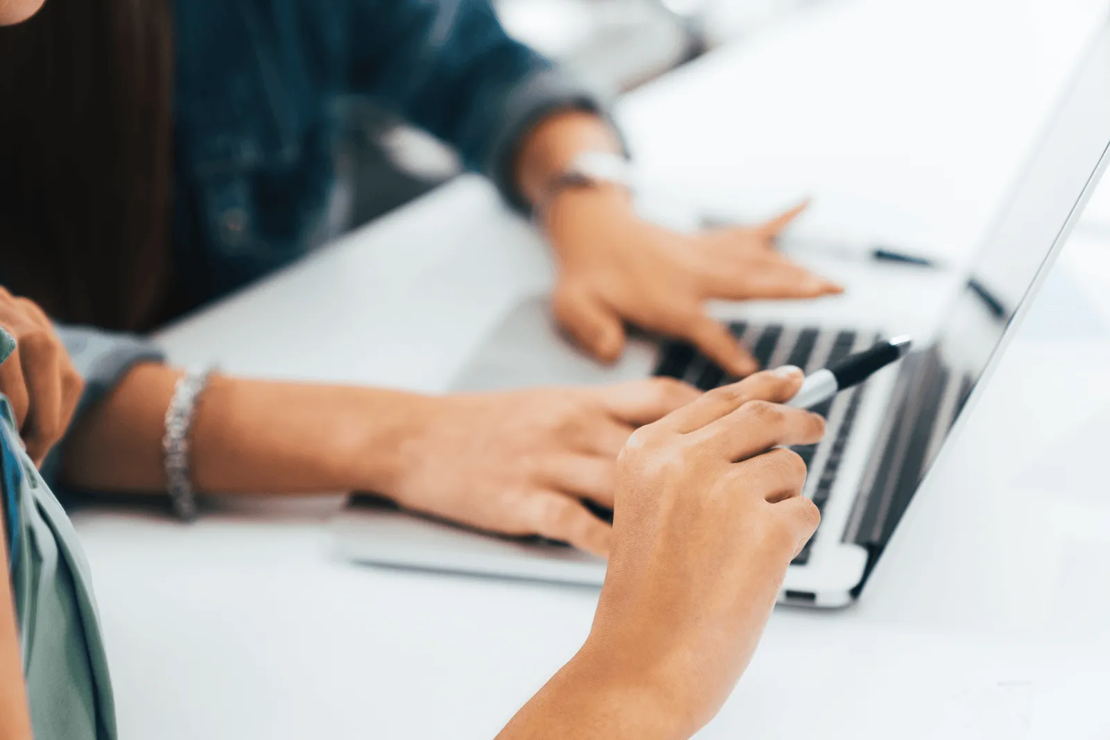 Close-up of two people collaborating using a laptop, one pointing at the screen with a pen and the other typing.