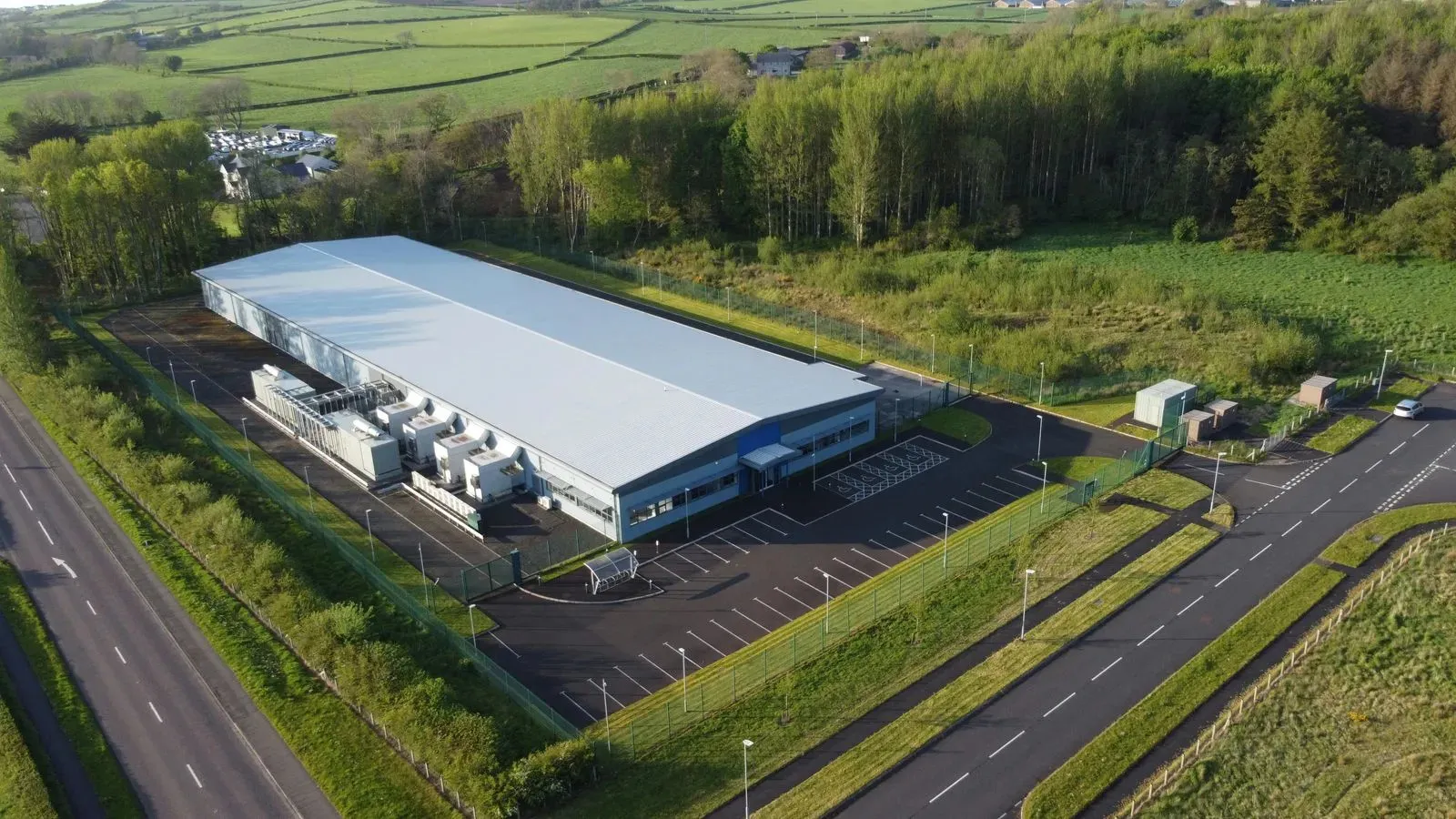 Aerial view of a large white industrial building with parking lots, surrounded by green fields and trees.