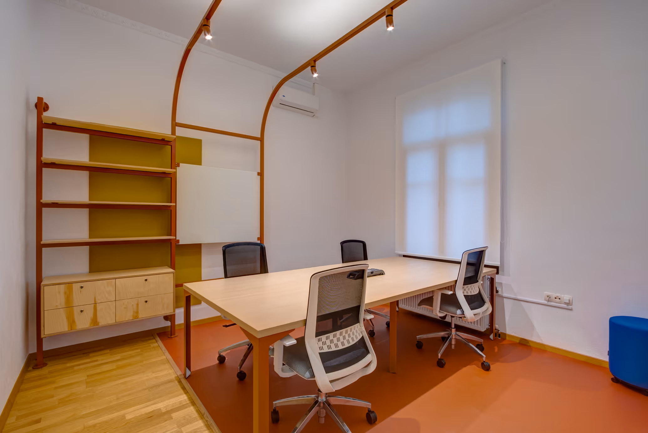 Interior meeting room with a large wooden table, four office chairs, built-in shelving, a whiteboard, and a window.