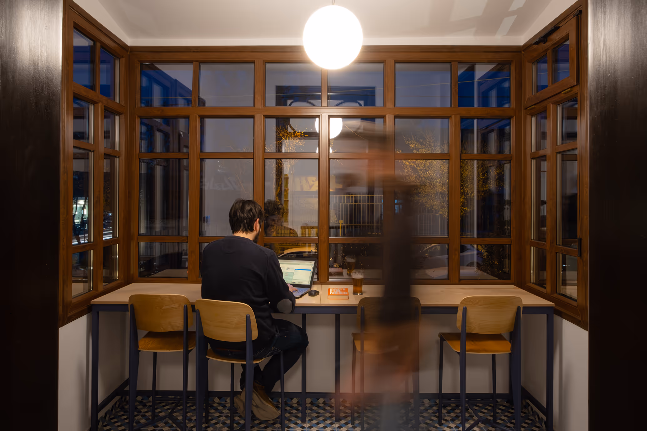 Person working on a laptop at a counter in a windowed seating area at night, with wooden chairs, a hanging light, and a blurred figure walking past.