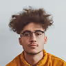 Portrait of a young man with curly hair, glasses, and a mustard yellow hoodie against a plain light background.