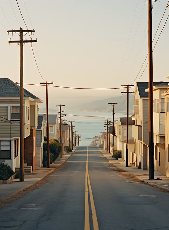 Empty street lined with houses and utility poles leading to a view of the ocean and distant hills under a clear sky.