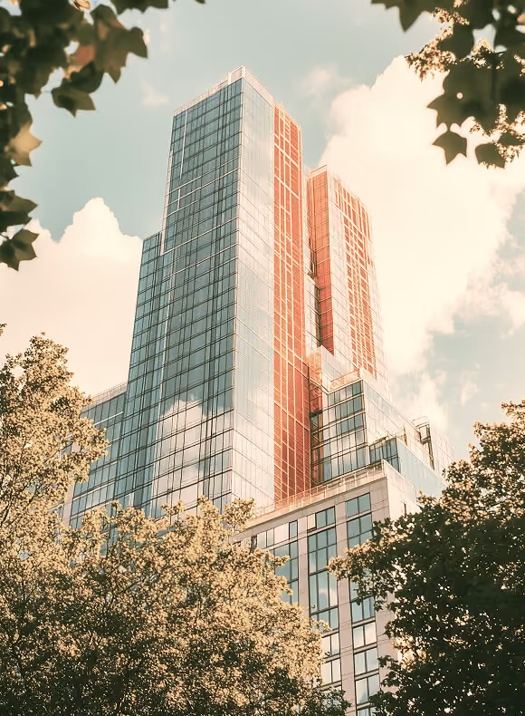 Modern glass skyscraper with reflections, framed by tree branches with green leaves under a cloudy sky.