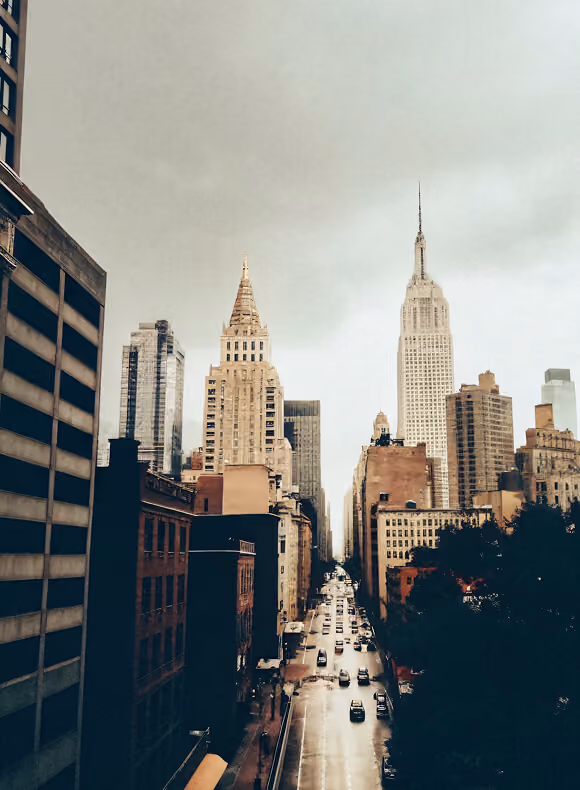 View of a busy New York City street with the Empire State Building and Chrysler Building visible under a cloudy sky.