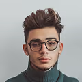 Portrait of a young man with dark styled hair, wearing round glasses and a green turtleneck sweater against a plain light background.