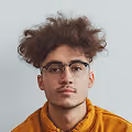 Young man with curly hair, glasses, and a brown jacket against a plain light background.