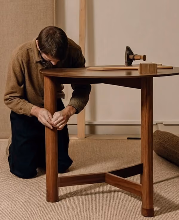 Man assembling a round wooden table, tightening the leg joint while kneeling on a carpeted floor.