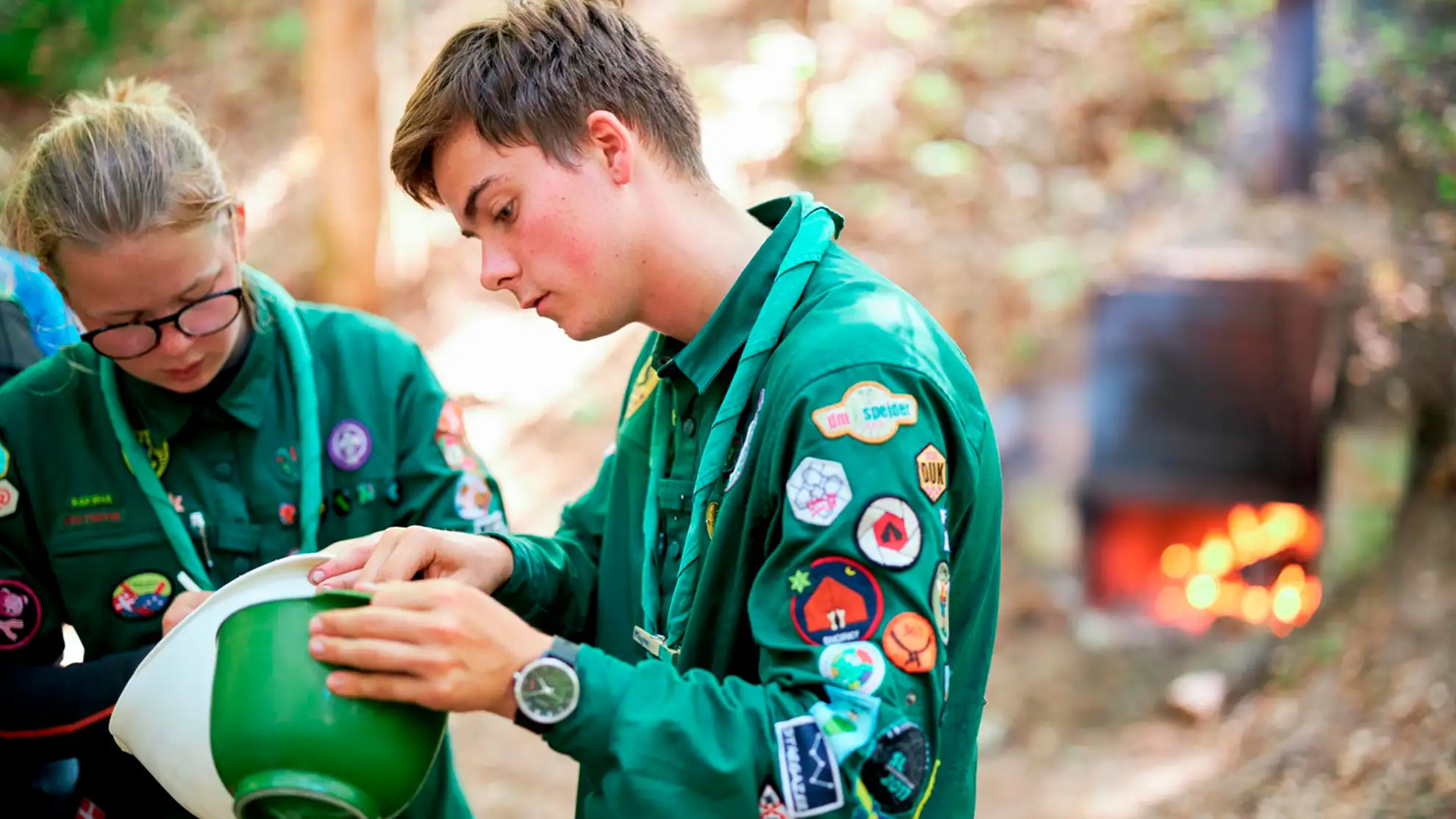 A girl and a boy scout standing with bowls in front of a fire