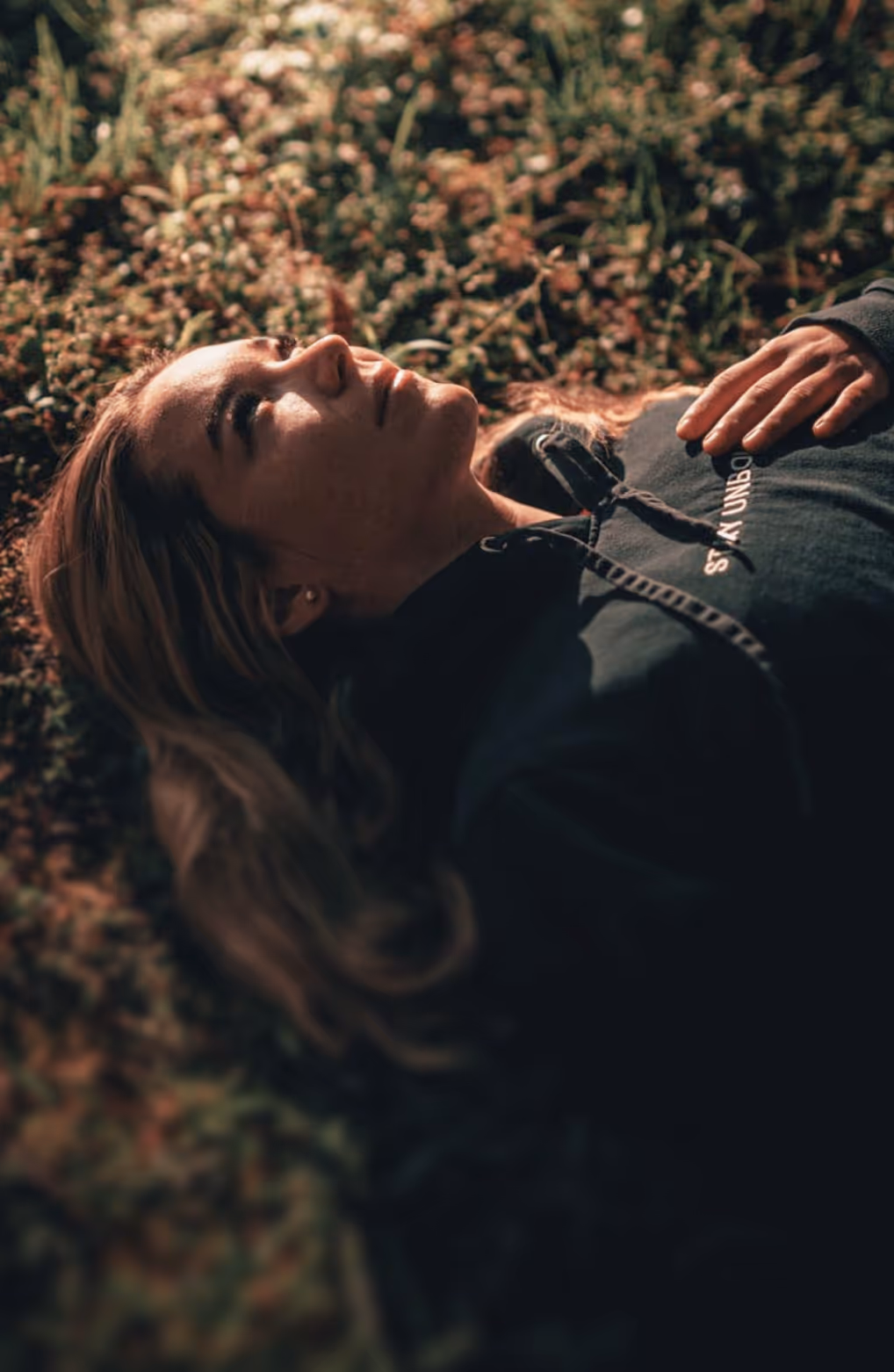 Young woman lying on grass with eyes open, wearing a dark hoodie in soft natural light.