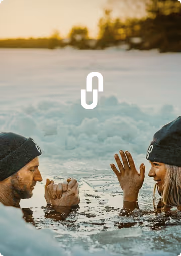 Two people wearing black beanies engage in an arm wrestling match while partially submerged in icy water surrounded by snow.