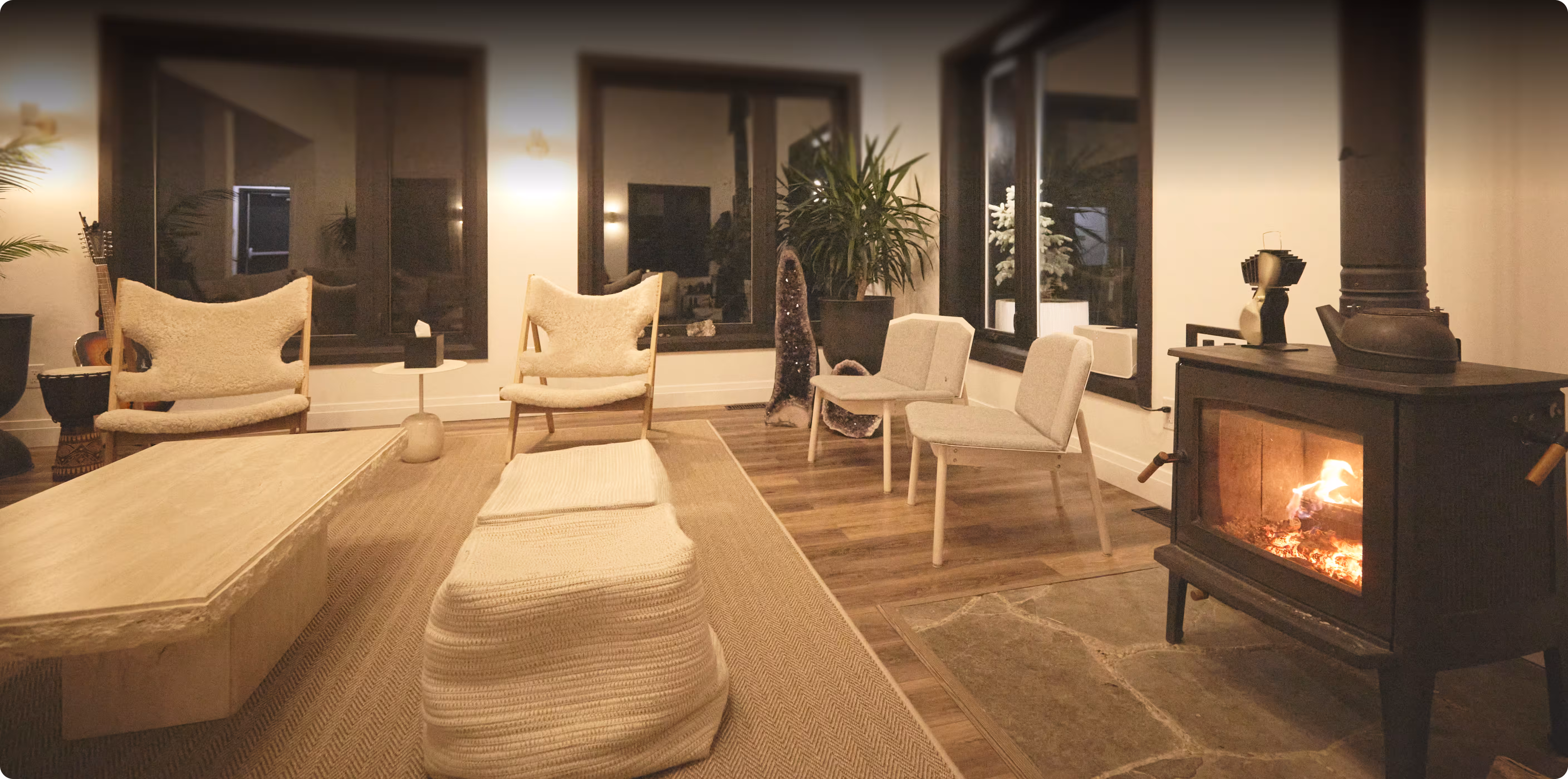 Cozy living room with a lit wood stove, two cushioned armchairs, two modern chairs, a knitted ottoman, wooden coffee table, and large windows.