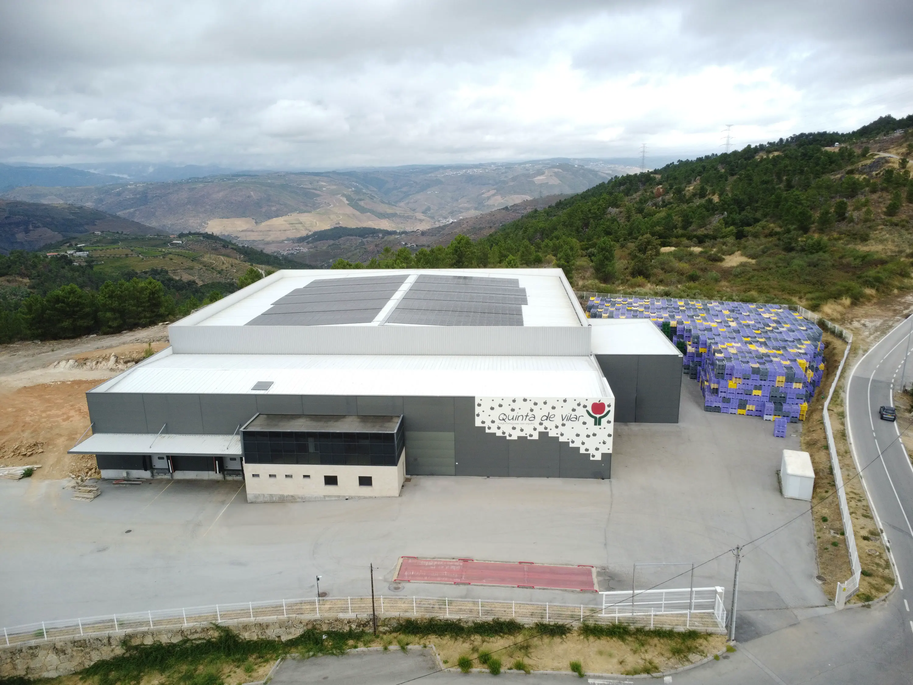 Edifício industrial cinzento com painéis solares no telhado e caixas empilhadas azuis e amarelas ao lado, situado numa área montanhosa.