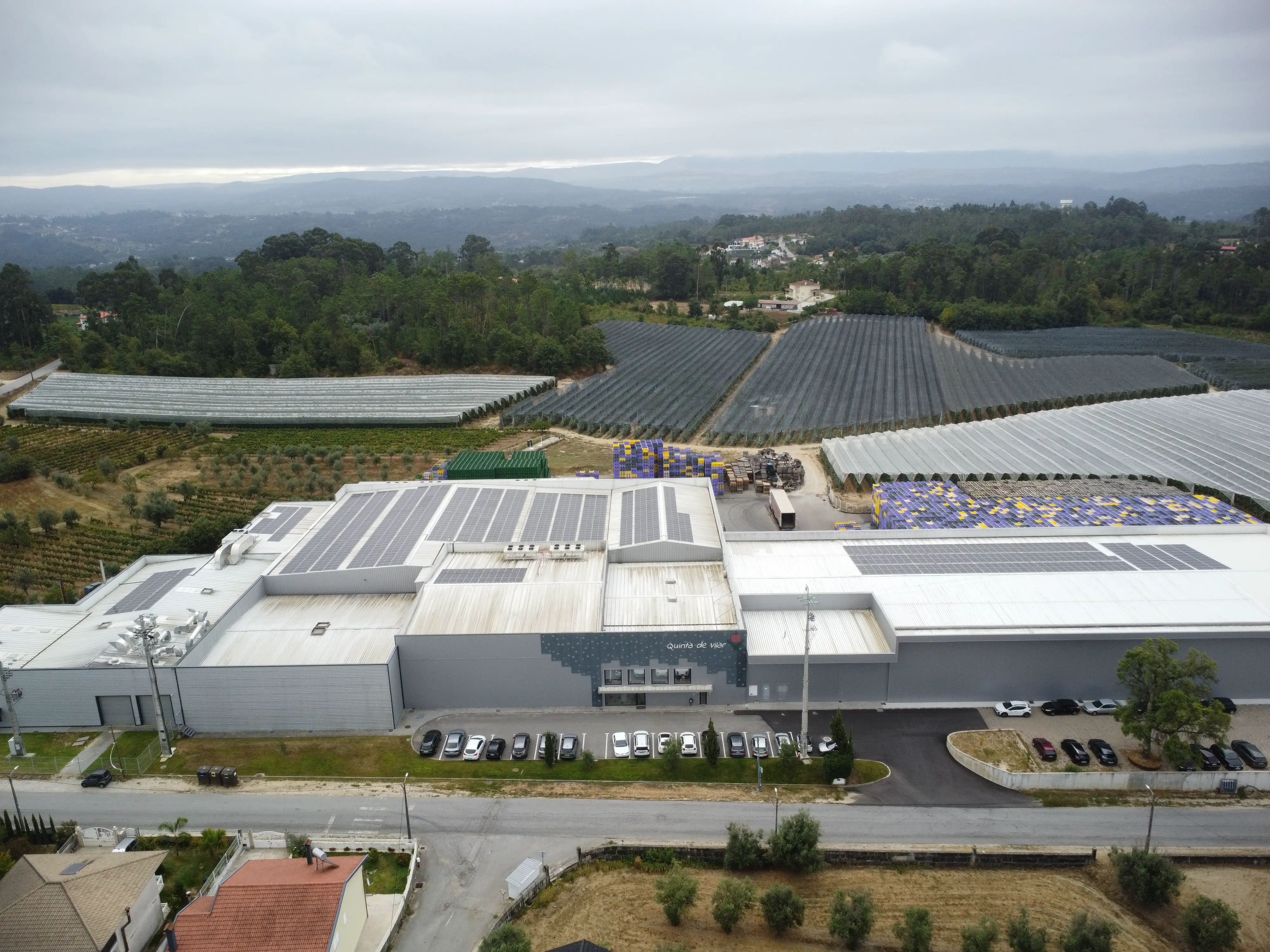 Vista aérea de uma grande instalação industrial com painéis solares no telhado, rodeada por áreas agrícolas cobertas por estufas e florestas ao fundo.