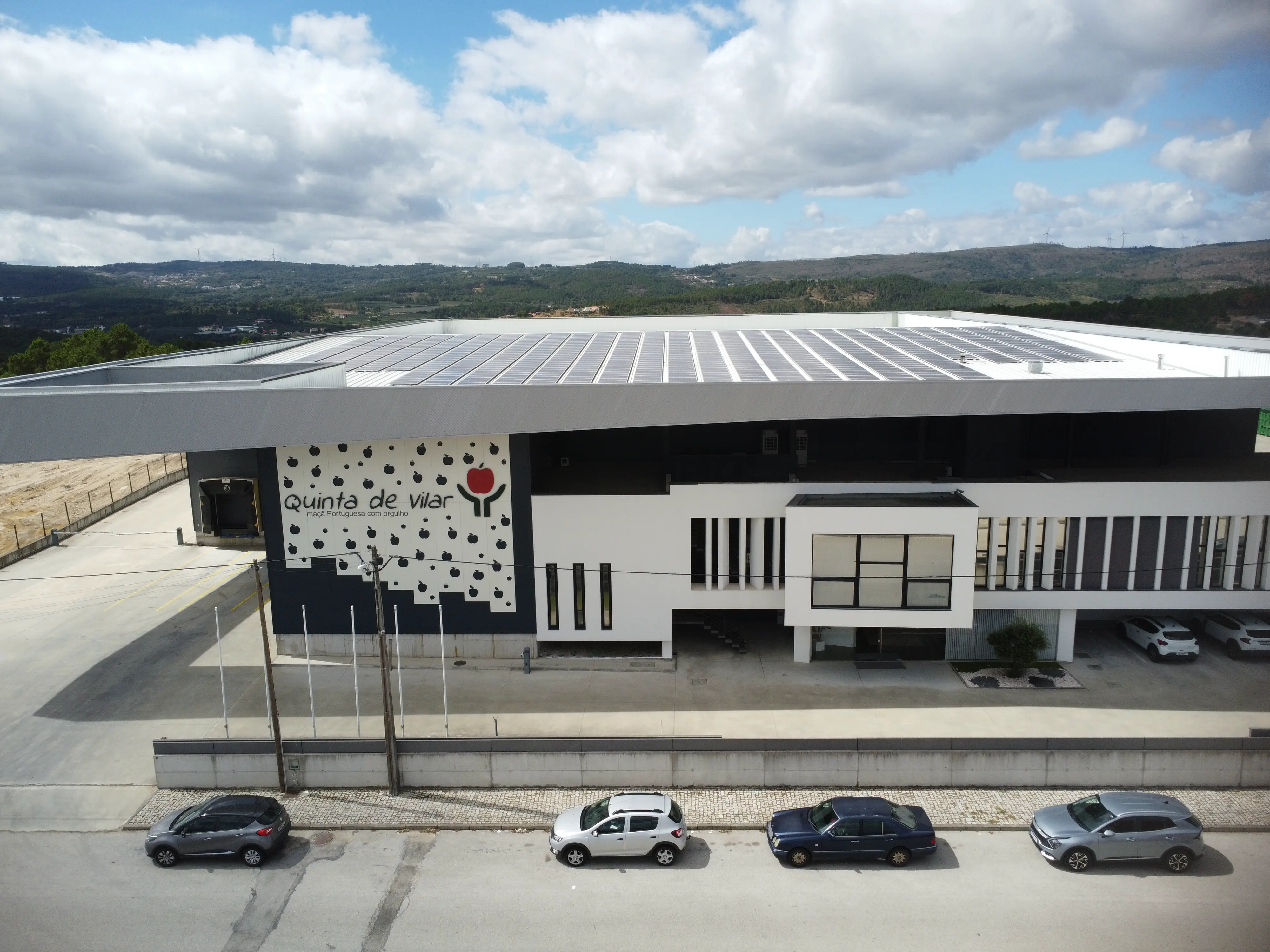 Vista aérea do edifício moderno da Quinta de Vilar com painéis solares no telhado e carros estacionados à frente.