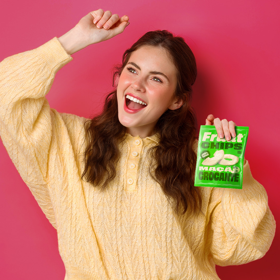 Mulher sorridente com suéter amarelo segurando um pacote verde de Fruut Chips sabor maçã crocante, fundo rosa.