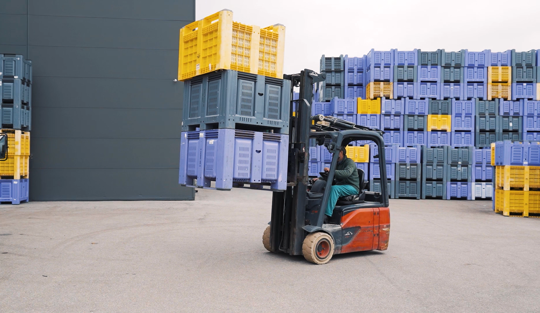 Edifício industrial moderno com painéis solares no telhado rodeado por caixas roxas empilhadas e vinhas verdes em paisagem montanhosa.
