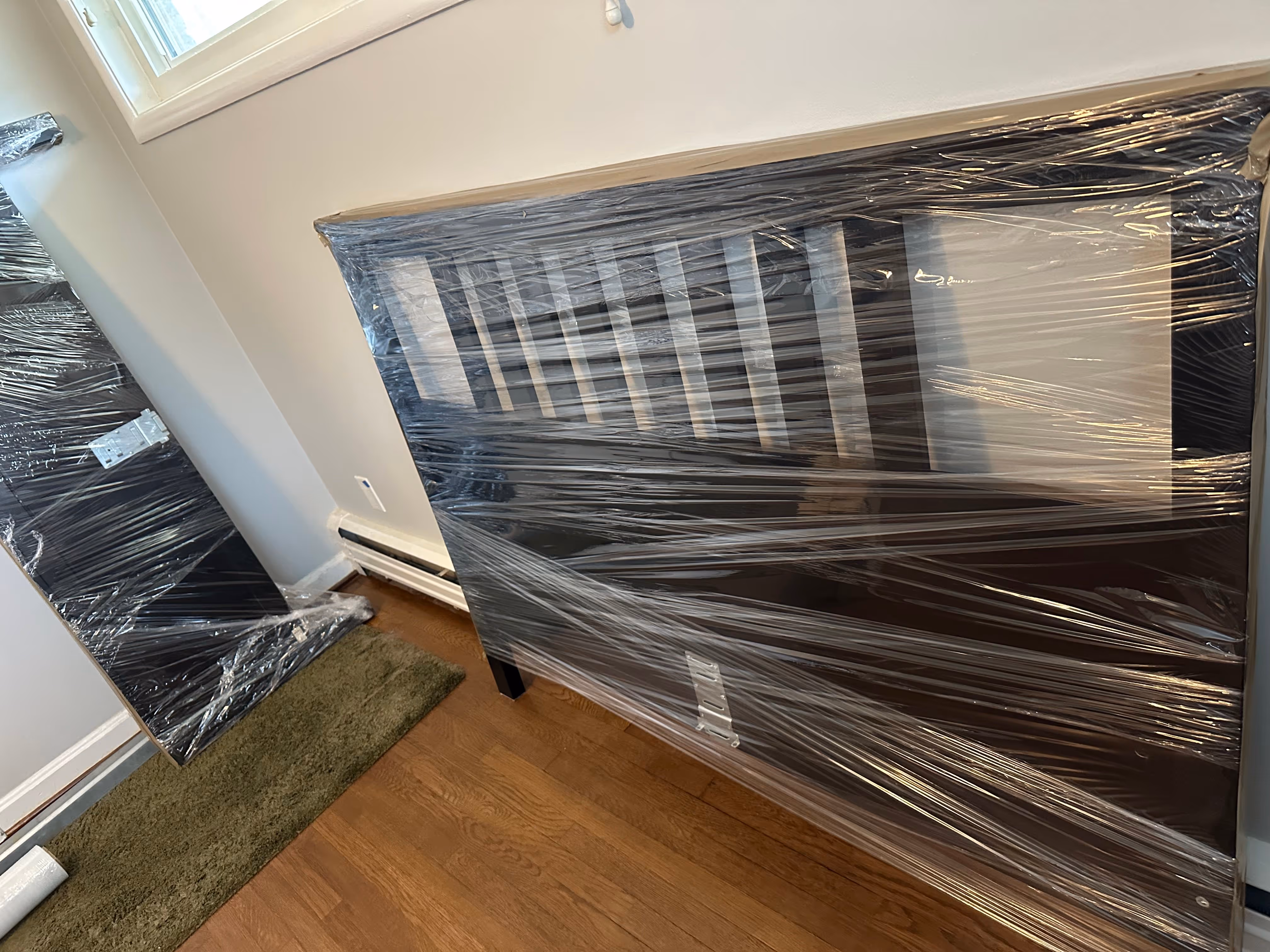 Two black wooden headboards wrapped in clear protective plastic leaning against a light-colored wall near a window.