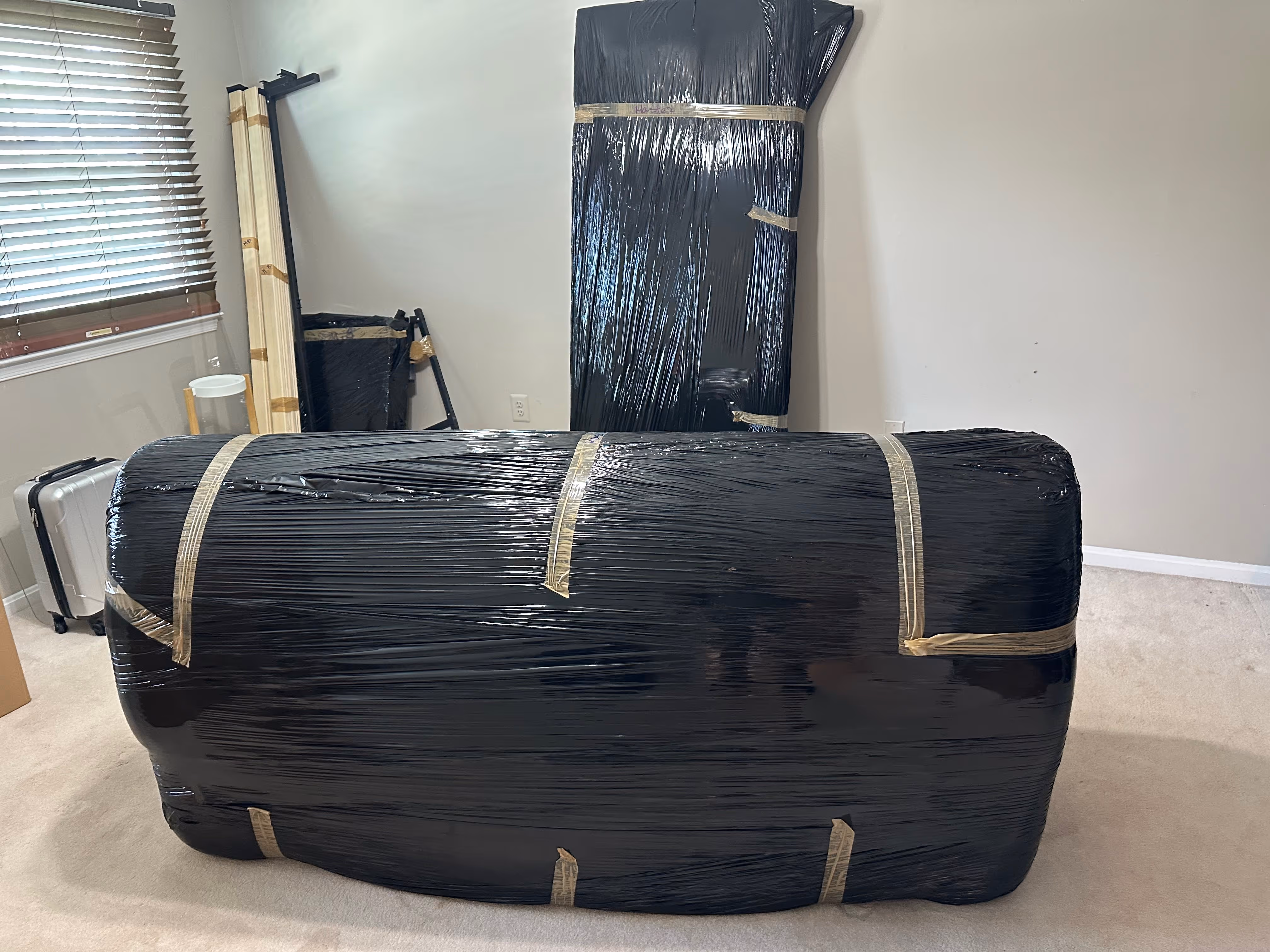 Two large furniture pieces wrapped in black plastic and secured with tape, placed in a room with beige carpet and window blinds.