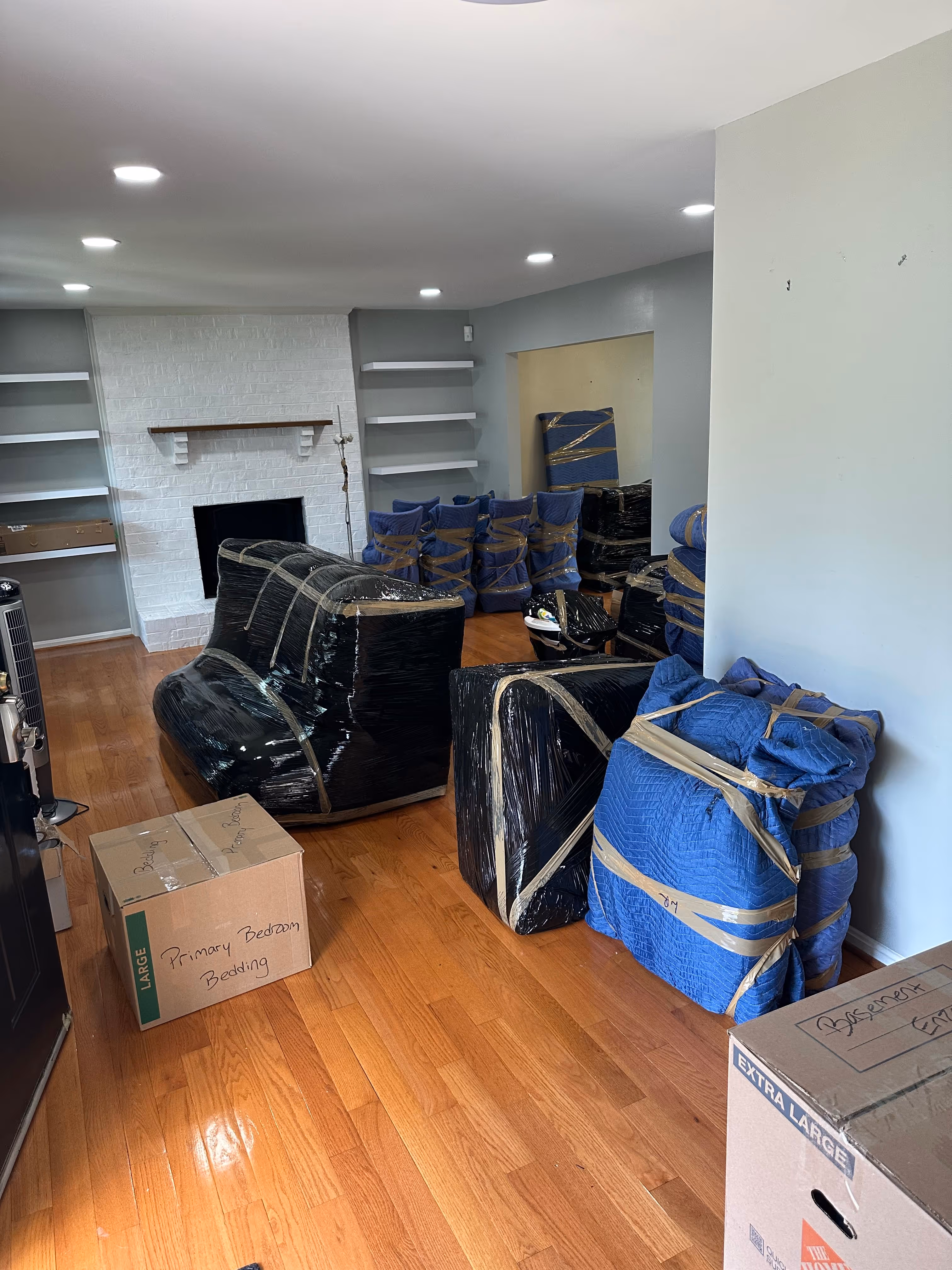 Living room with furniture wrapped in plastic and tape for moving, along with labeled moving boxes on wooden floor.
