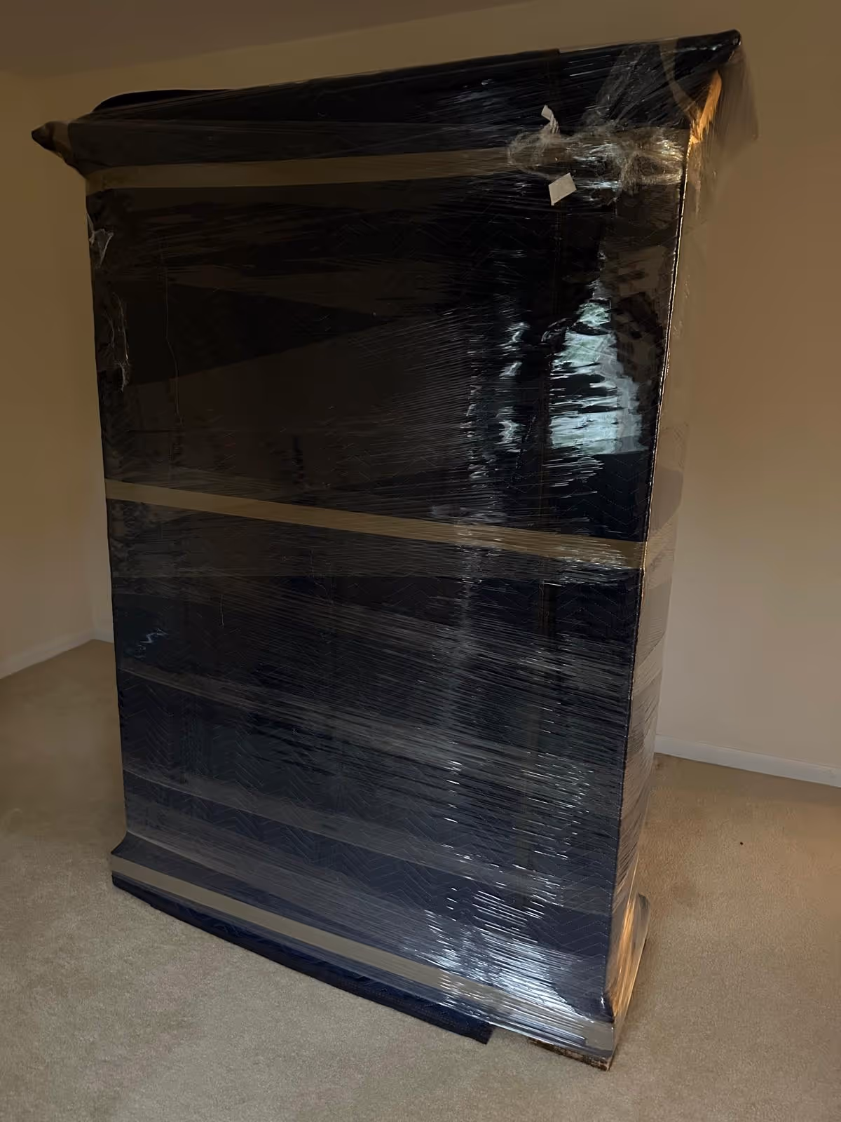 Large piece of furniture wrapped in black protective moving blanket and covered with clear plastic wrap standing on carpeted floor.