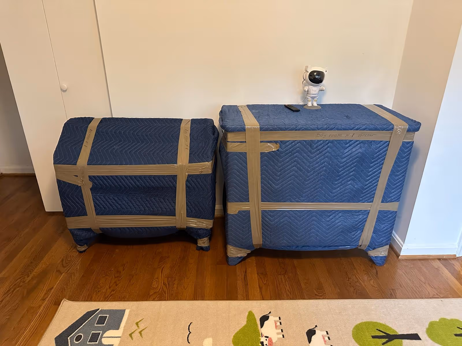 Two pieces of furniture wrapped in blue moving blankets and secured with brown packing tape, one topped with a small astronaut figurine and a TV remote.