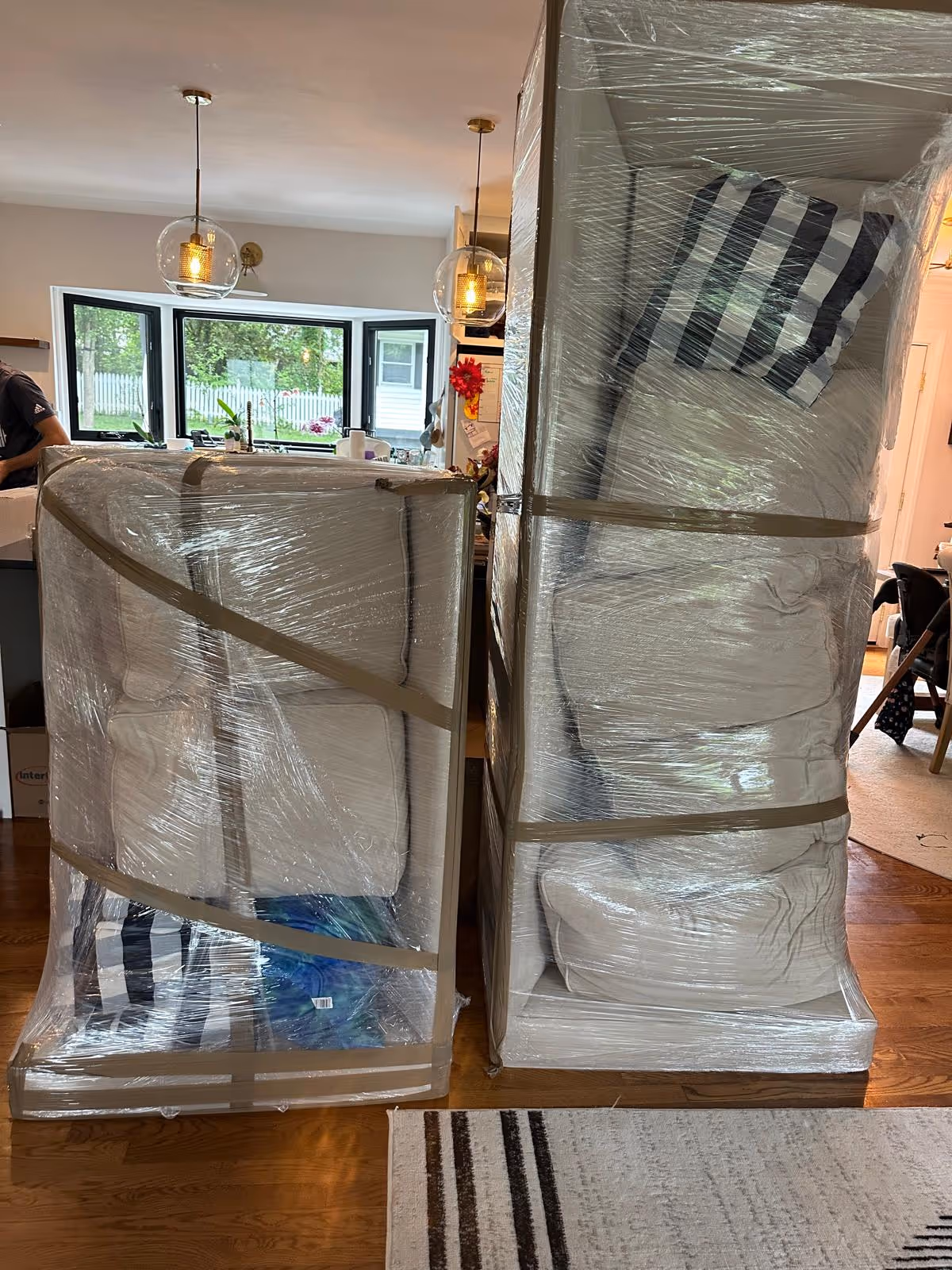 Two white couches wrapped in plastic wrap and secured with tape inside a home with hardwood floors and pendant lights.