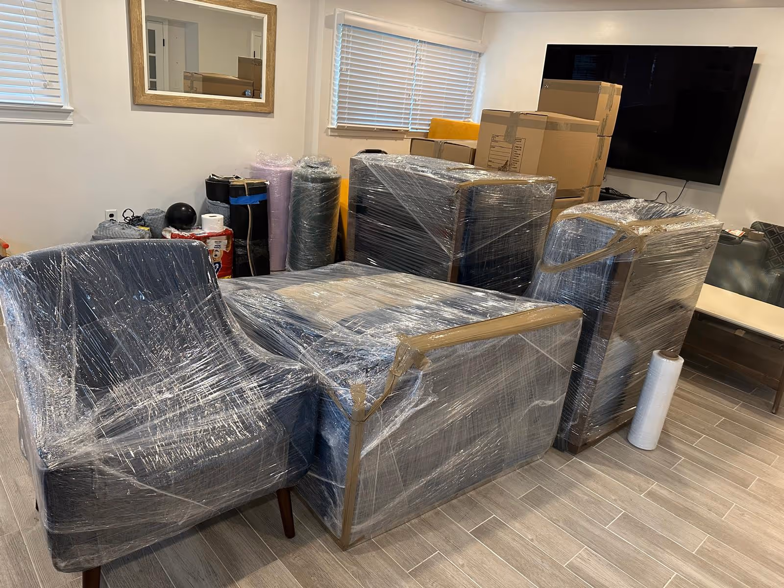 Living room furniture including armchairs and ottomans wrapped in plastic and tape, with stacked cardboard boxes and rolled items in the background.