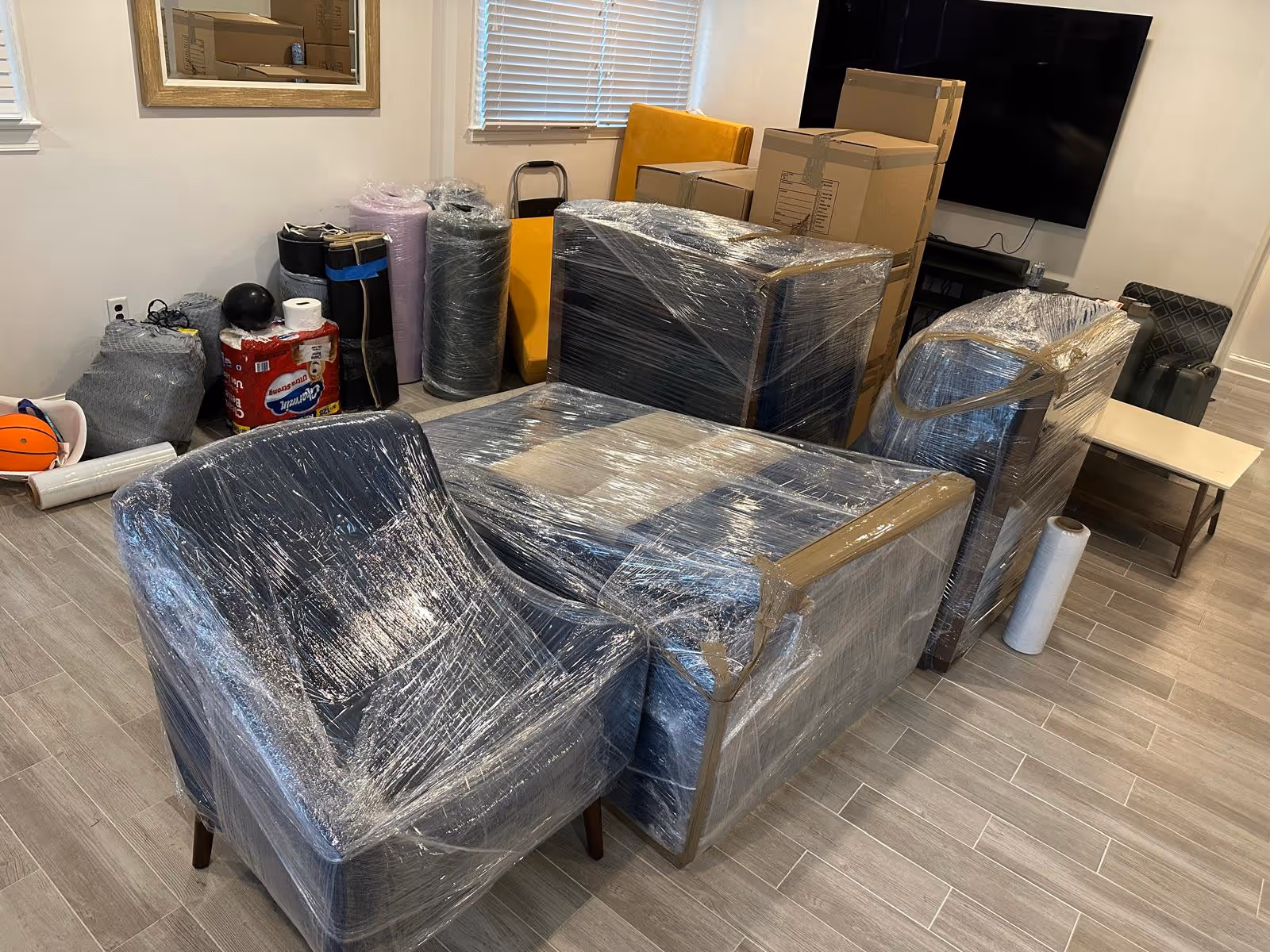 Living room furniture wrapped in plastic wrap ready for moving, with packed boxes and household items in the background.