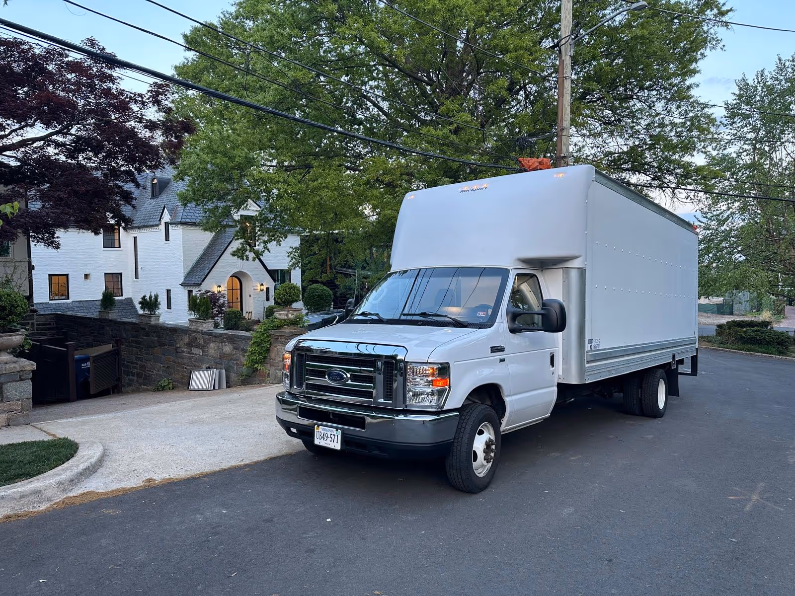 White Ford box truck parked on a residential street in front of a large white house with greenery and trees.