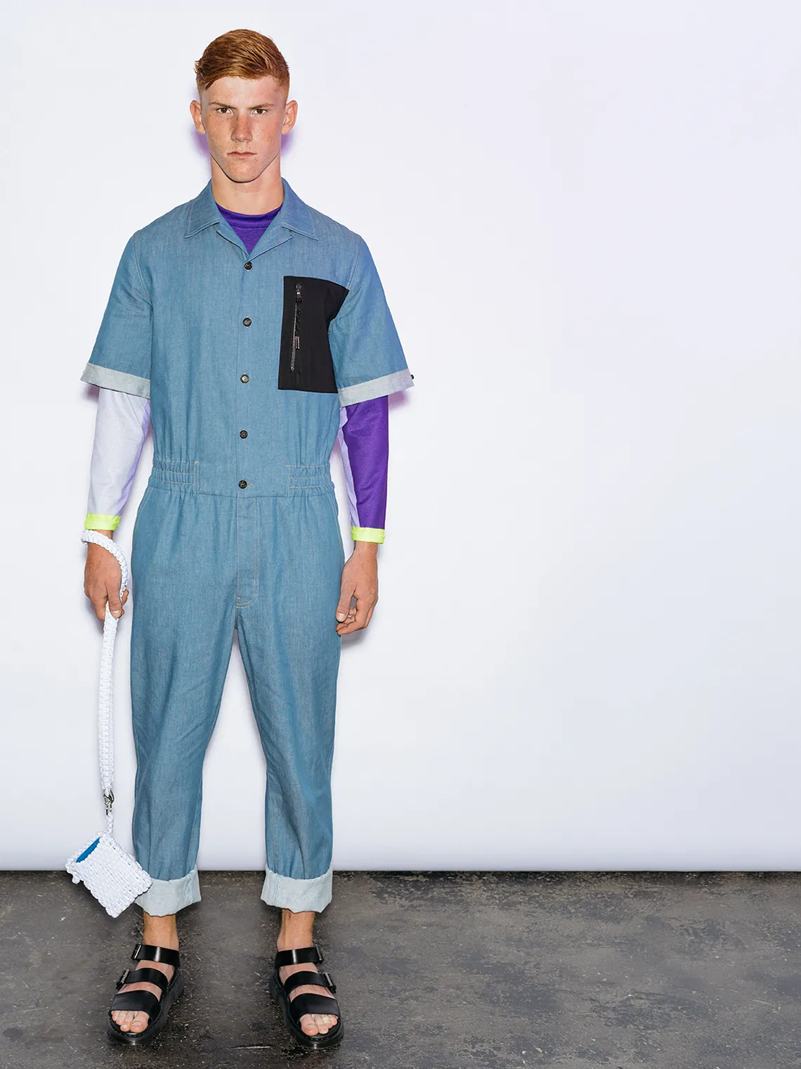 Model wearing the PSEUDONYM Spring Summer 2020 light blue technical denim jumpsuit with a black zip pocket, layered over a purple and lilac compression tee, styled with black sandals and a white paracord accessory.