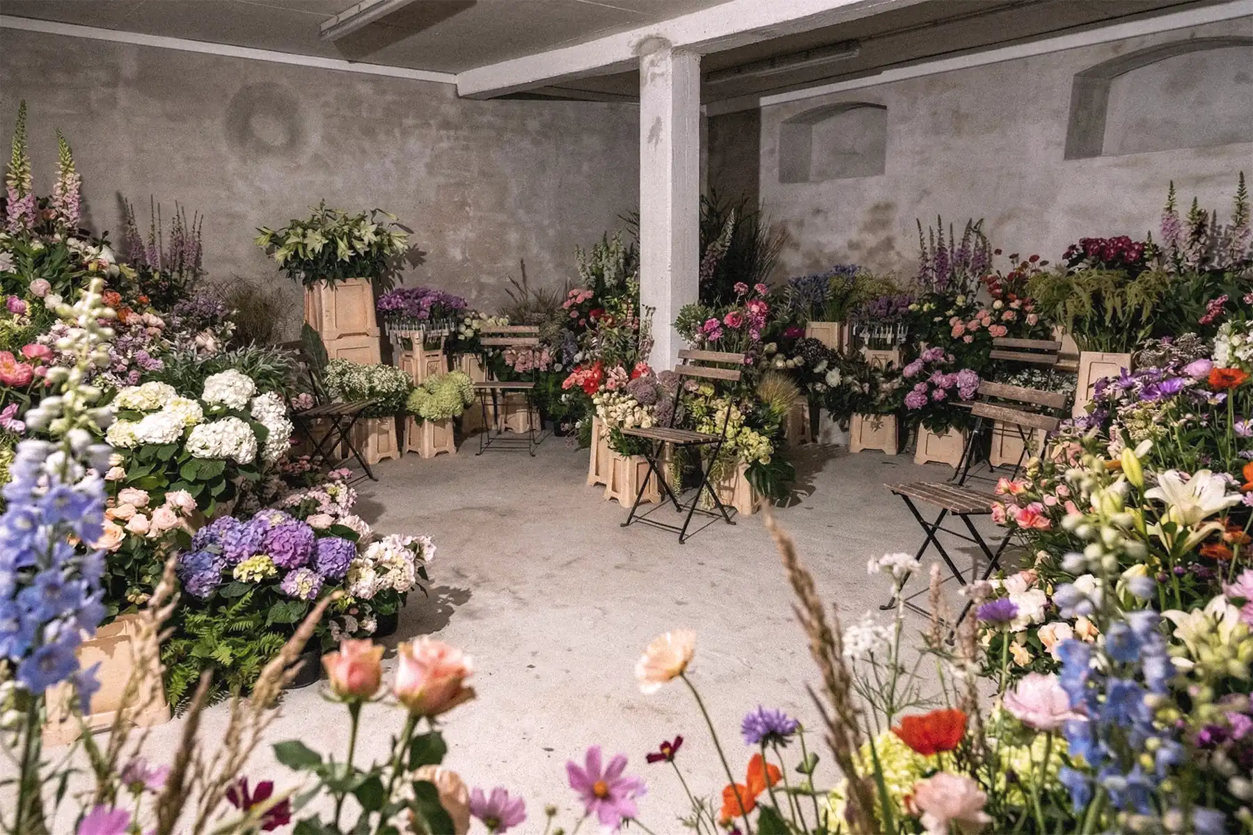 PSEUDONYM Spring Summer 2020 show production floral installation with abundant flowers, café chairs, and a raw concrete interior arranged as an immersive presentation space.