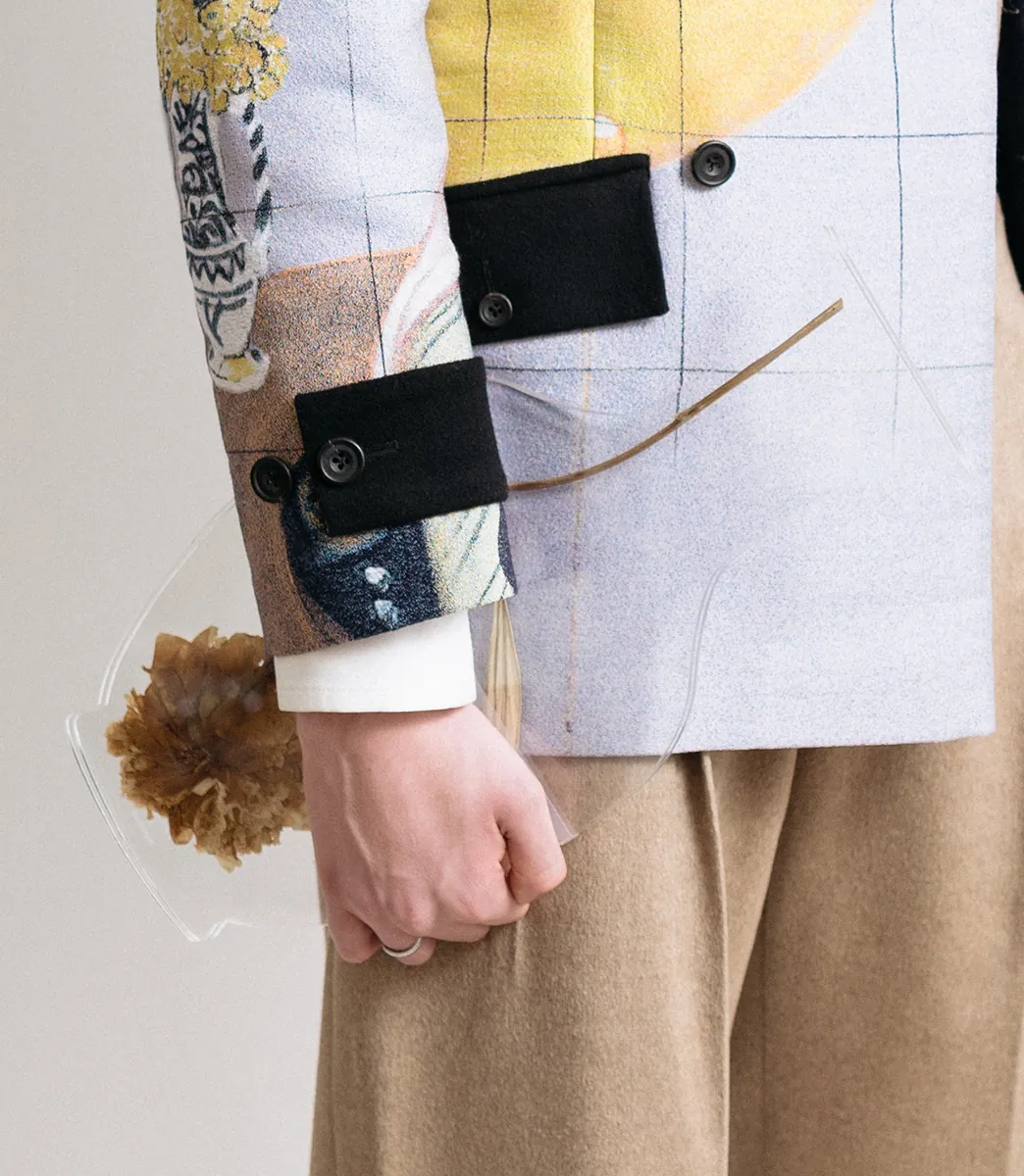 Close-up of a tailored jacquard jacket with contrast turnback cuffs and a clear plexiglass vase clutch encasing dried flowers from PSEUDONYM Autumn Winter 2019.