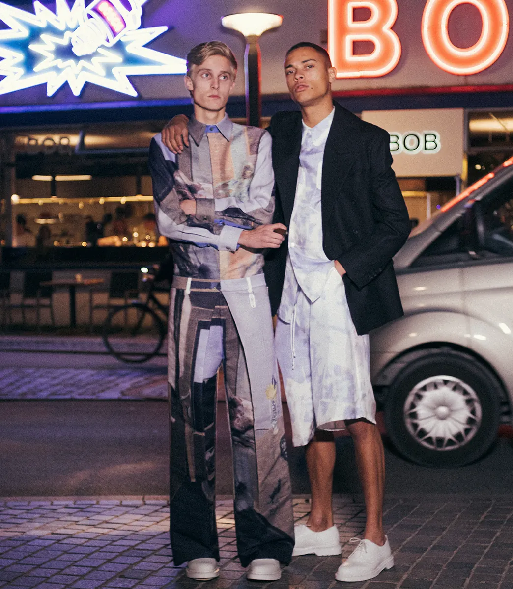Two models wearing PSEUDONYM Autumn Winter 2019 against neon signage, featuring a jacquard shirt with pleated trousers and a tailored jacket over a printed silk twill shirt and shorts.